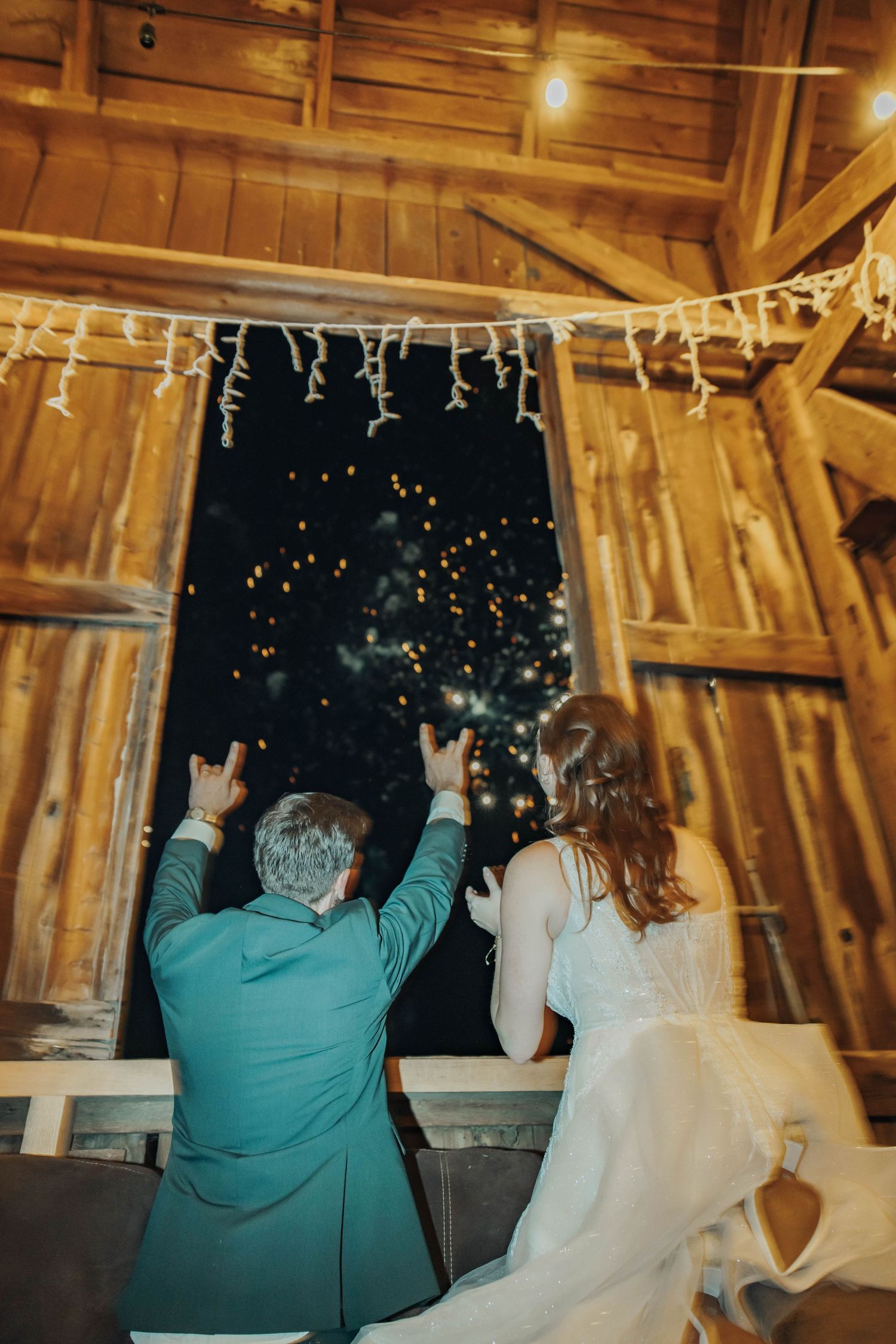 Couple watches fireworks display through barn window with twinkling lights overhead.