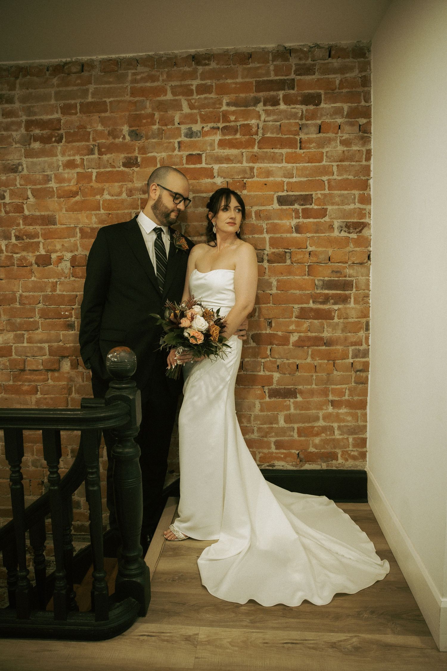 Bride in white strapless gown holds bouquet beside groom in black suit against rustic brick wall.
