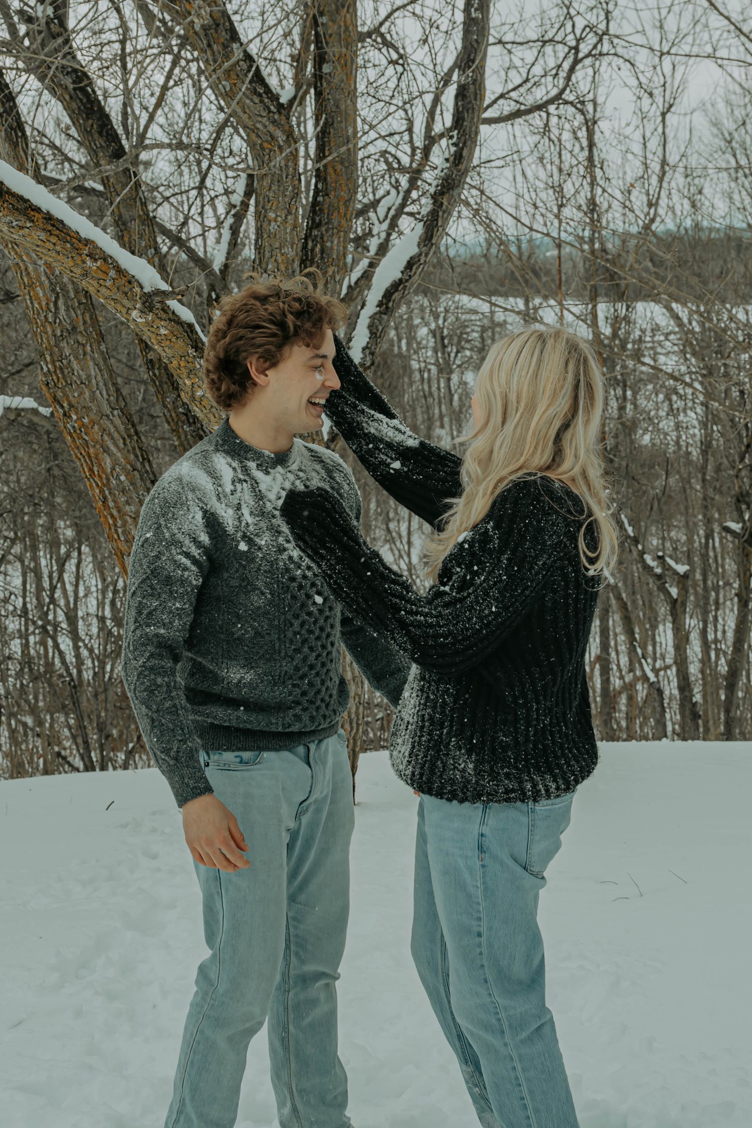 A couple in matching dark sweaters share a playful moment in the snow near a bare winter tree.