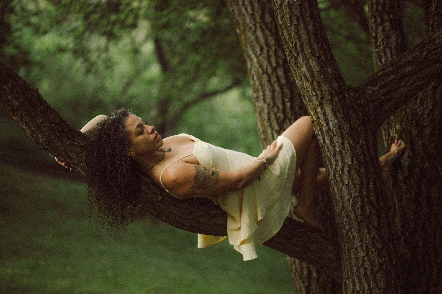 Woman with curly hair and tattoo resting peacefully on tree branches in a lush green forest wearing a white dress.