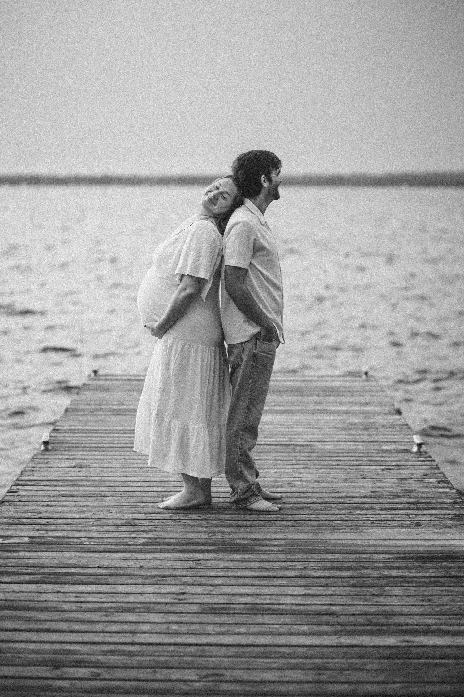 Black and white photo of a pregnant couple standing back-to-back on a wooden pier by the sea.