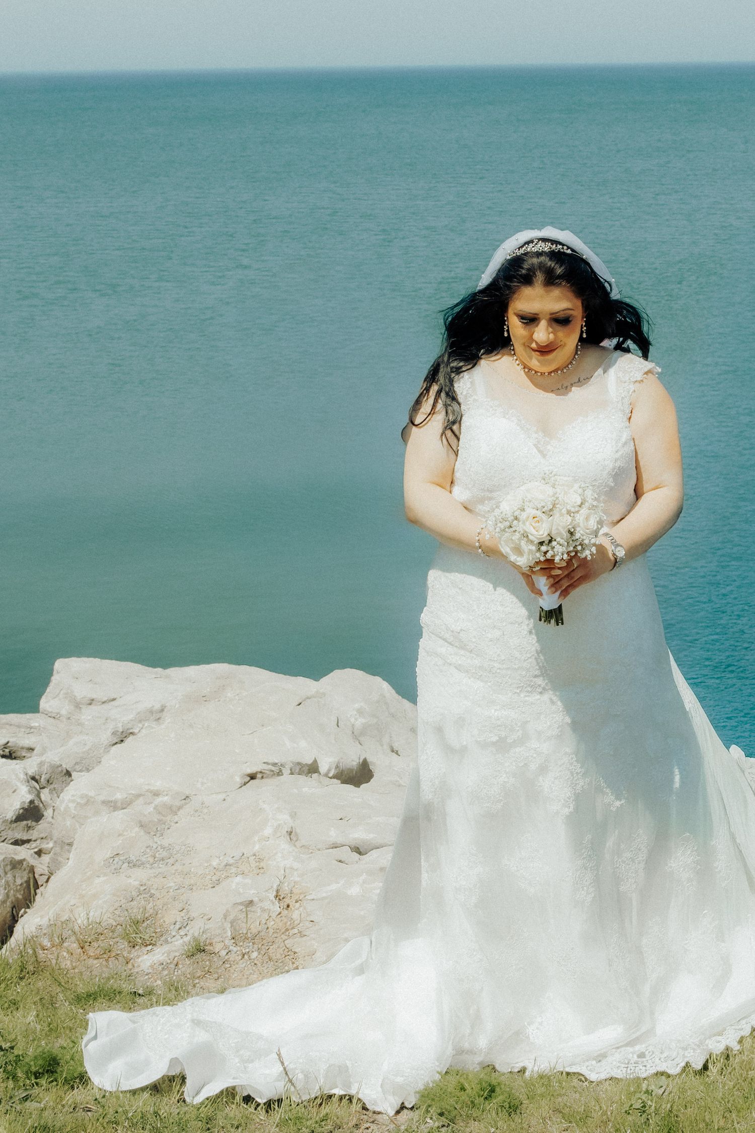 Bride in white lace gown holding bouquet on rocky seaside cliff with turquoise ocean backdrop.