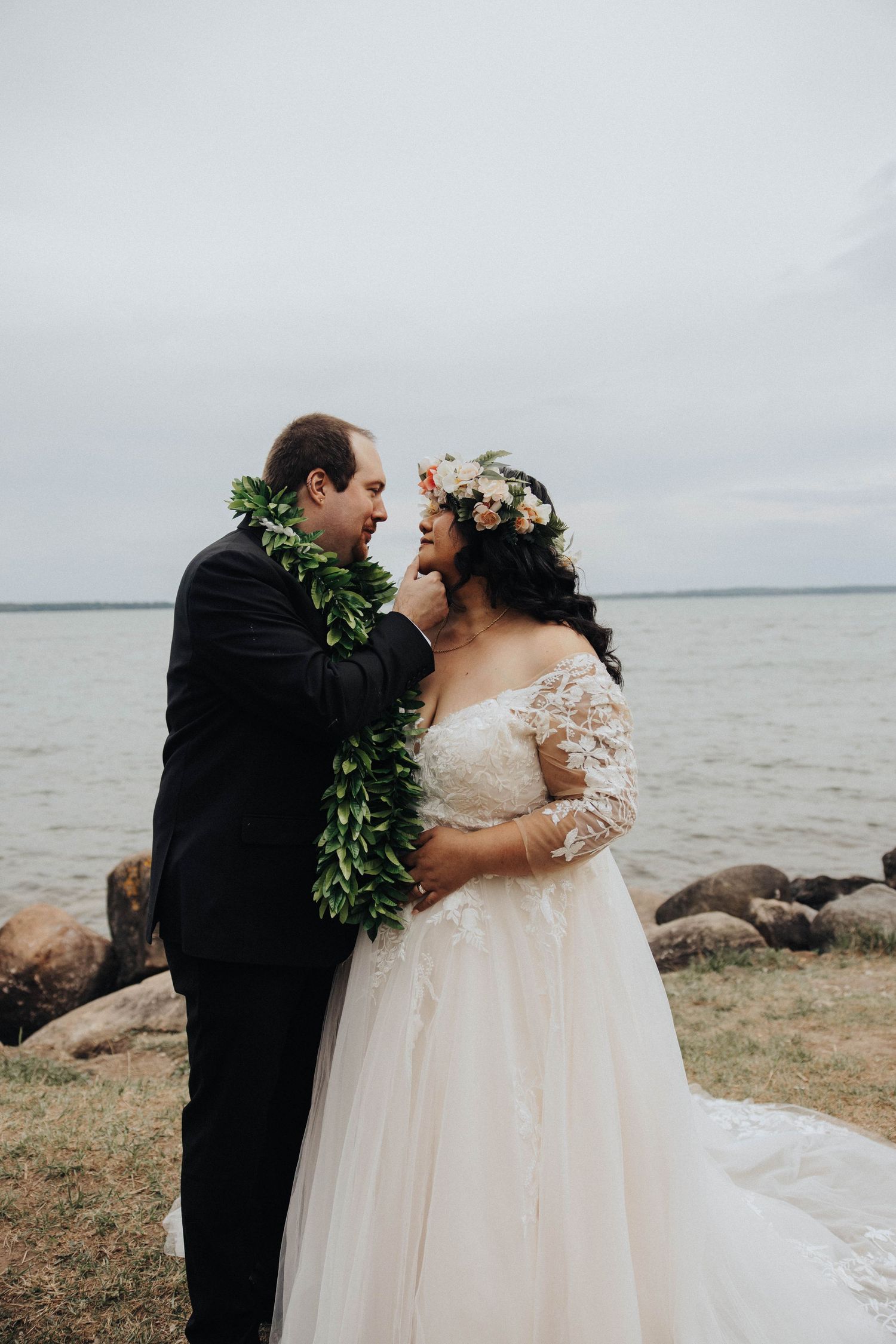 Romantic wedding portraits against a serene waterfront backdrop with rocks and cloudy skies.