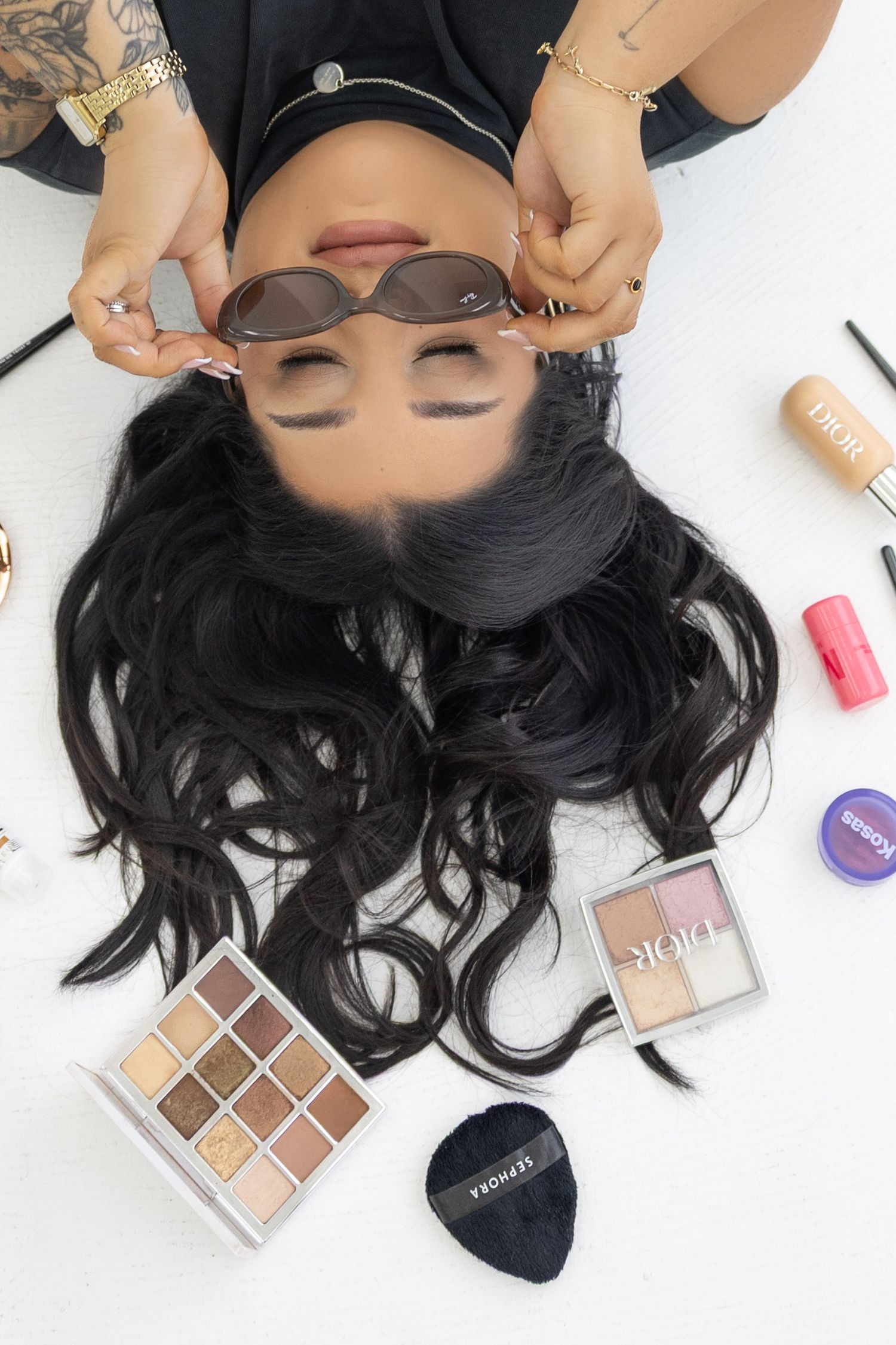 Woman lying down surrounded by Dior makeup, eyeshadow palettes, and beauty products, posing with sunglasses.