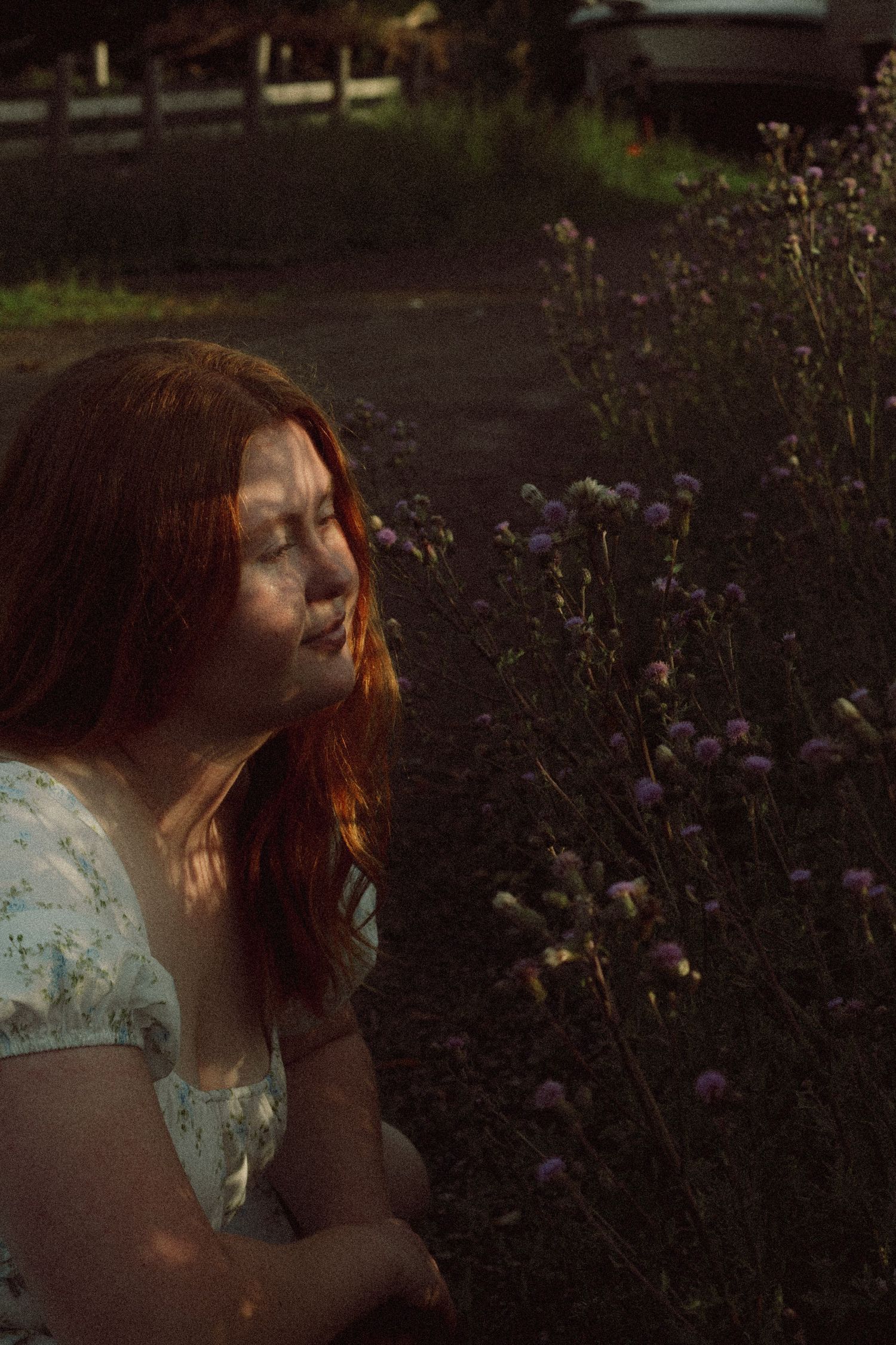 Young woman with red hair sits near wildflowers in golden hour light, wearing a floral dress outdoors.