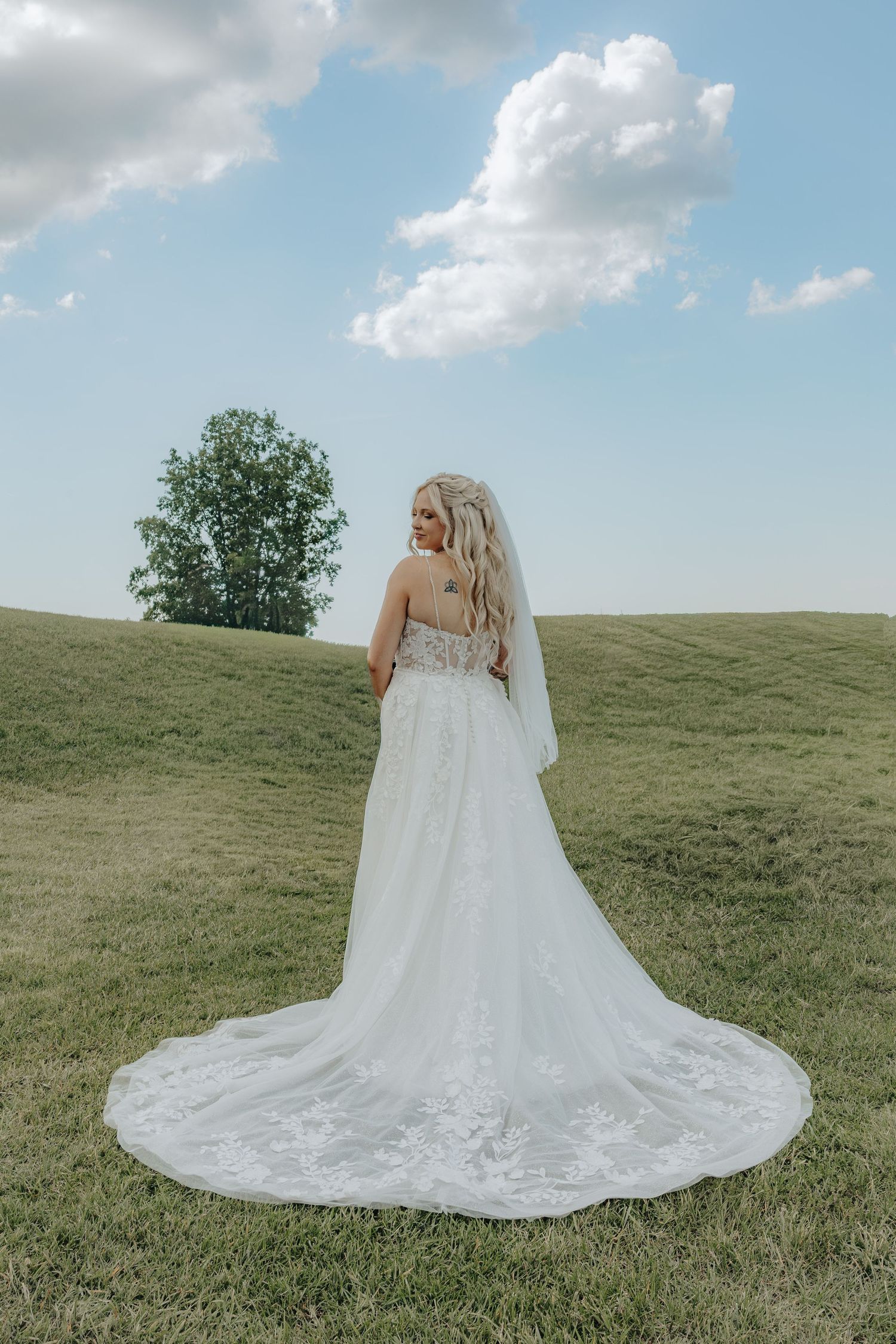Bride in white lace wedding gown with veil and long train stands on green hillside under blue sky.