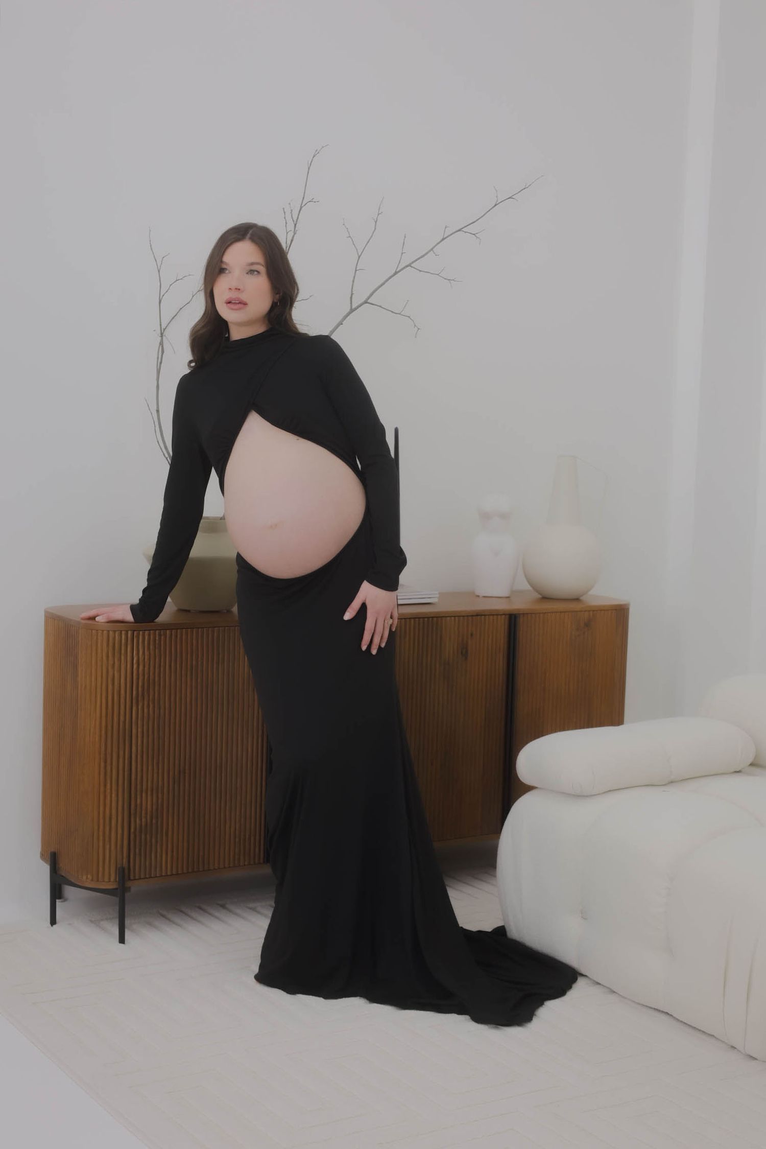 Dramatic maternity portrait in black dress beside vintage wooden cabinet in white room.