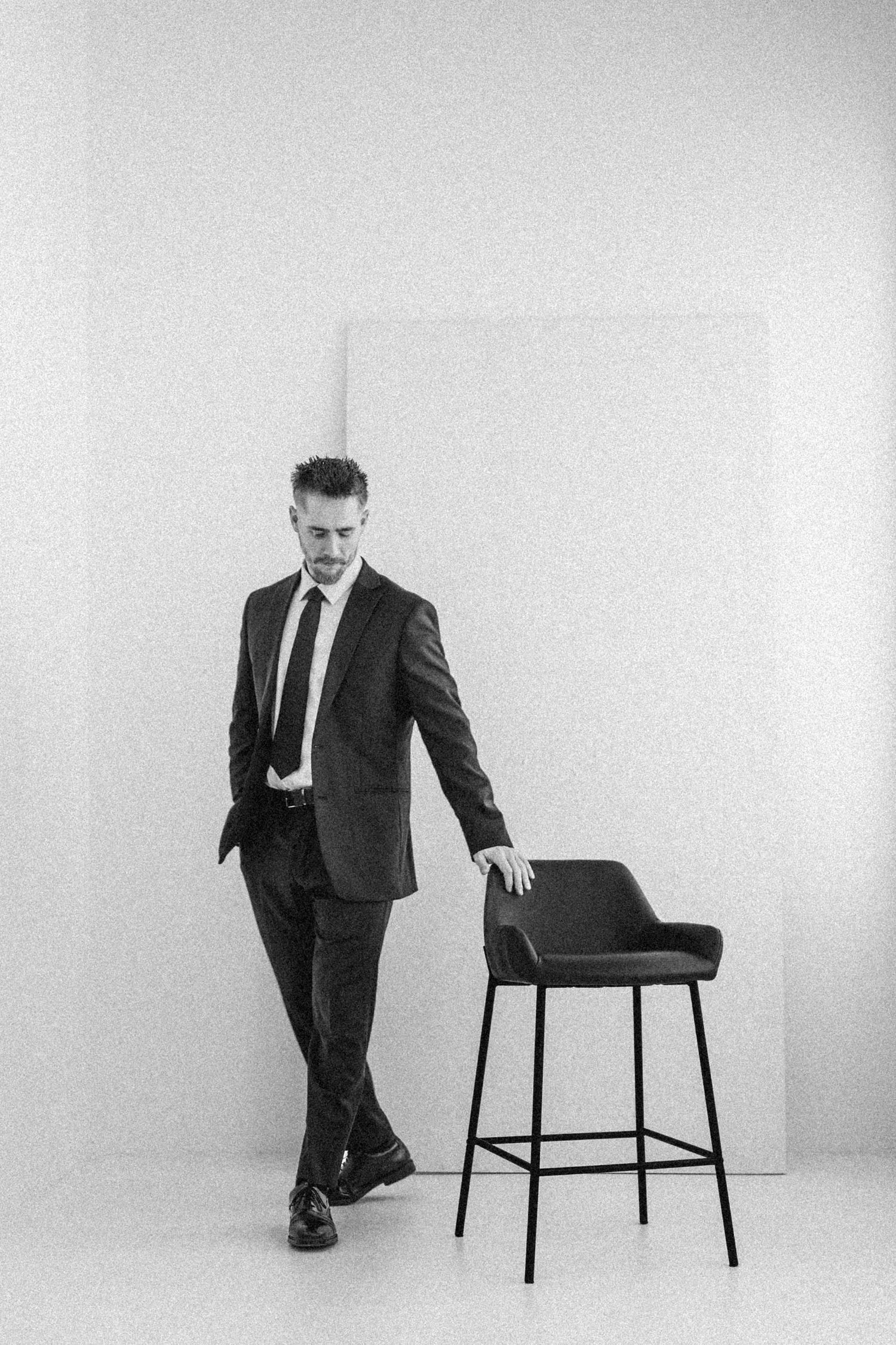 A black and white series showing a person in a suit interacting with a minimalist black chair against a white backdrop.