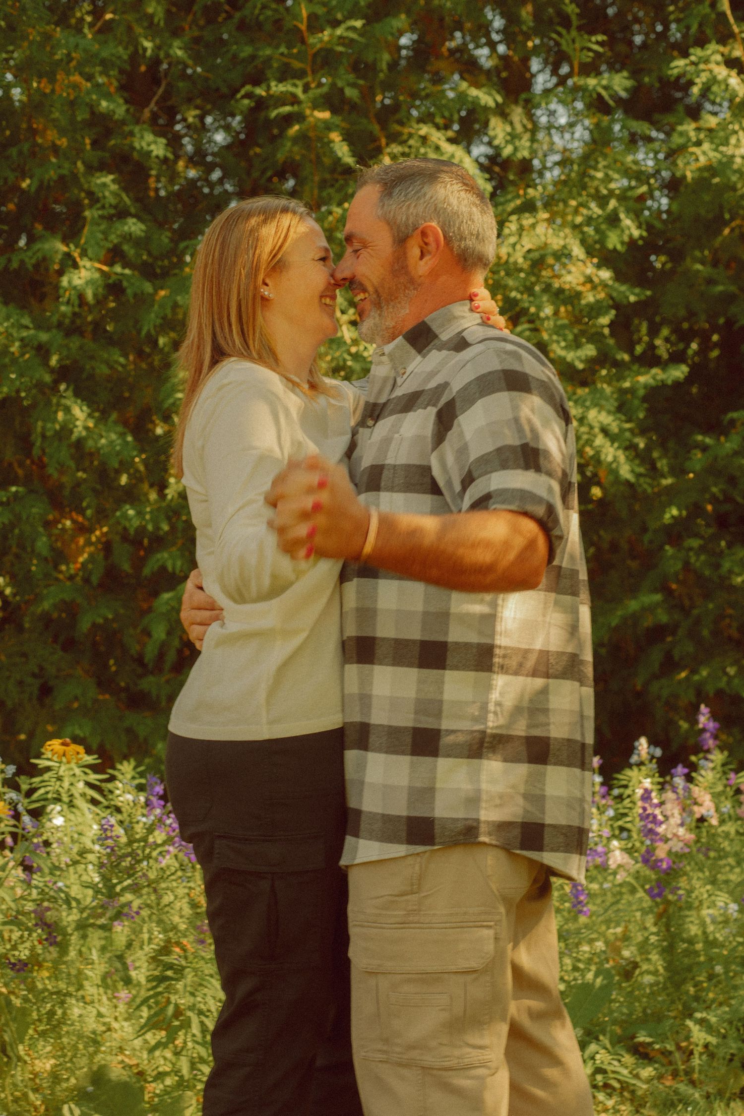 A couple embraces and smiles at each other in a lush garden setting with colorful flowers and green trees.