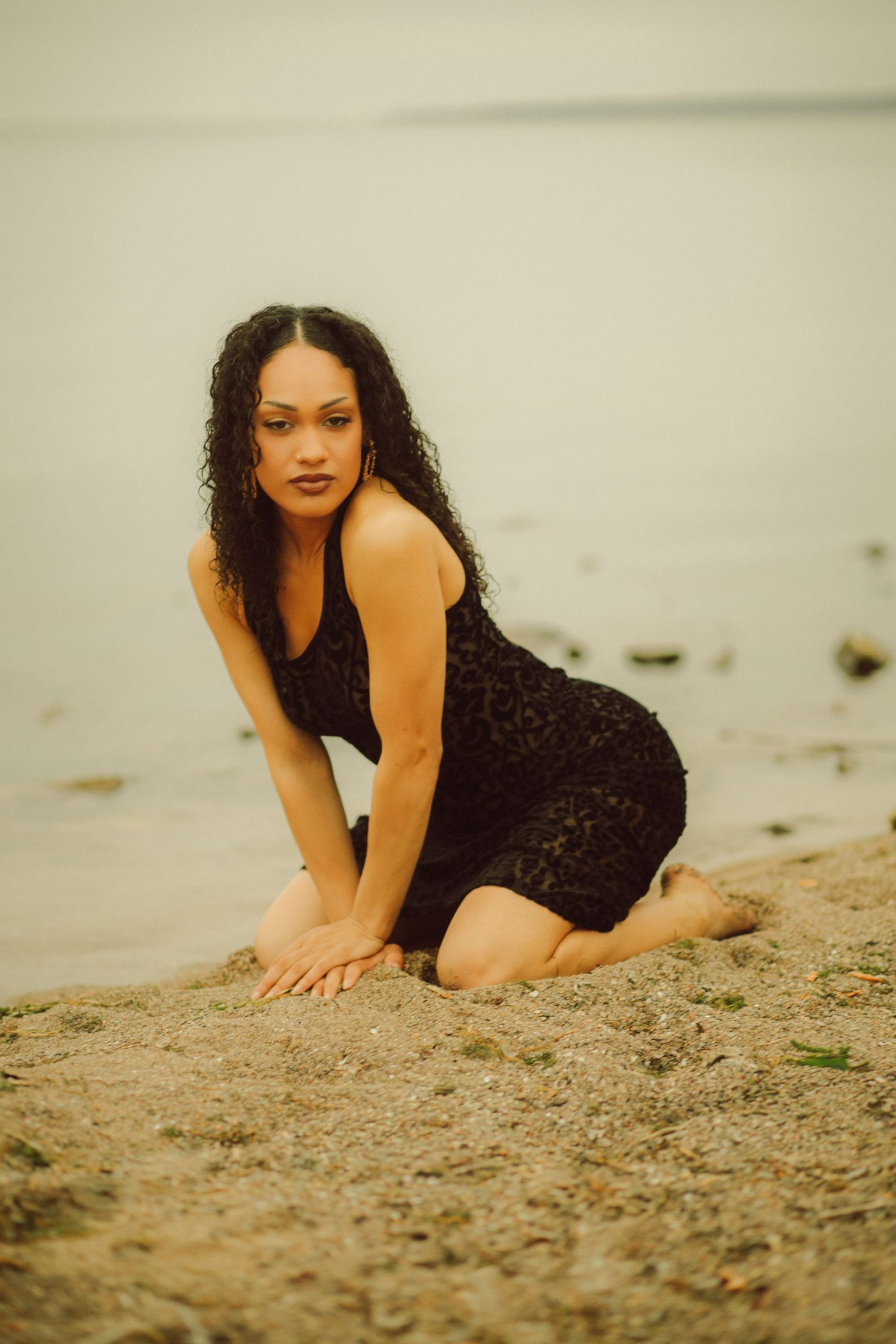 A woman with curly black hair in a black lace dress poses on sandy beach shore with calm water in background.