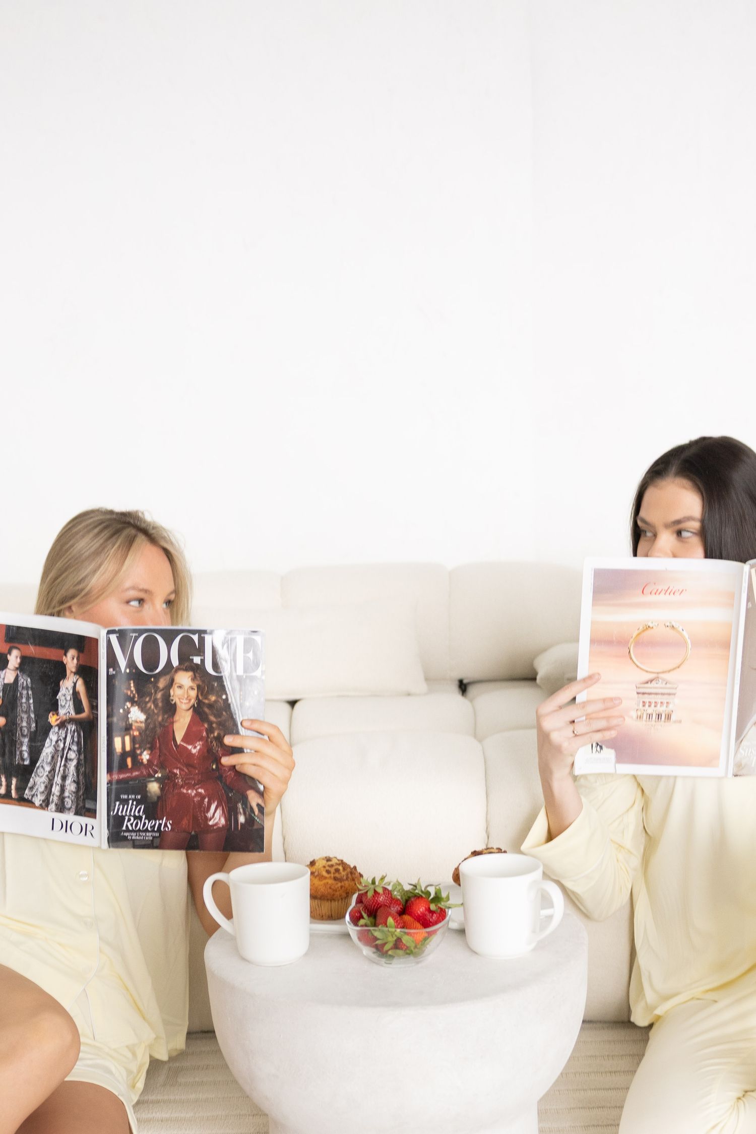 Two women reading Vogue magazines on white sofa with coffee and strawberries on round table in bright room.