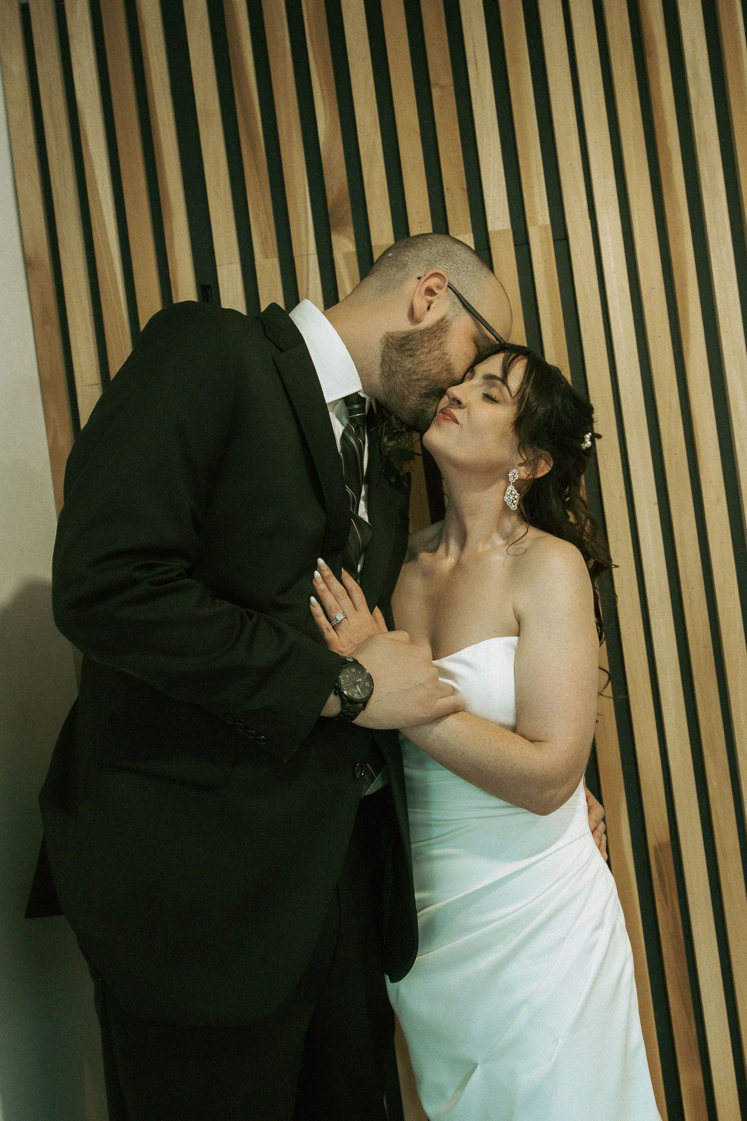 A groom in a black suit kisses his bride in a white strapless gown against a wooden slat wall.