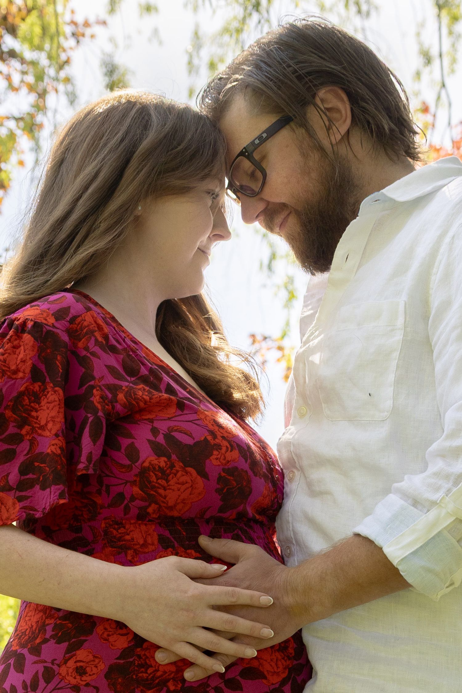 Pregnant couple tenderly touching baby bump outdoors, man in glasses and white shirt, woman in red floral dress, autumn trees background.