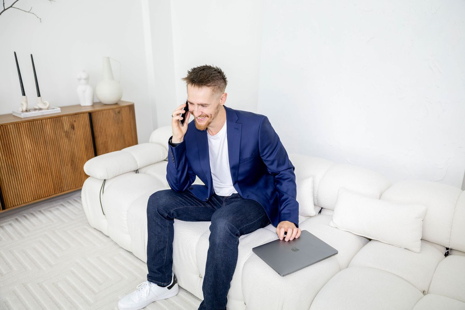 Professional in navy suit sits on white couch while working on laptop and talking on smartphone.
