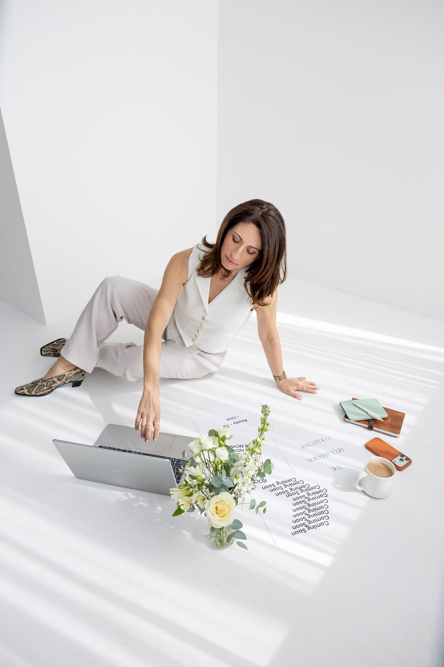 Woman in white outfit sitting on floor with laptop, flowers, notebook, and coffee cup in bright workspace.