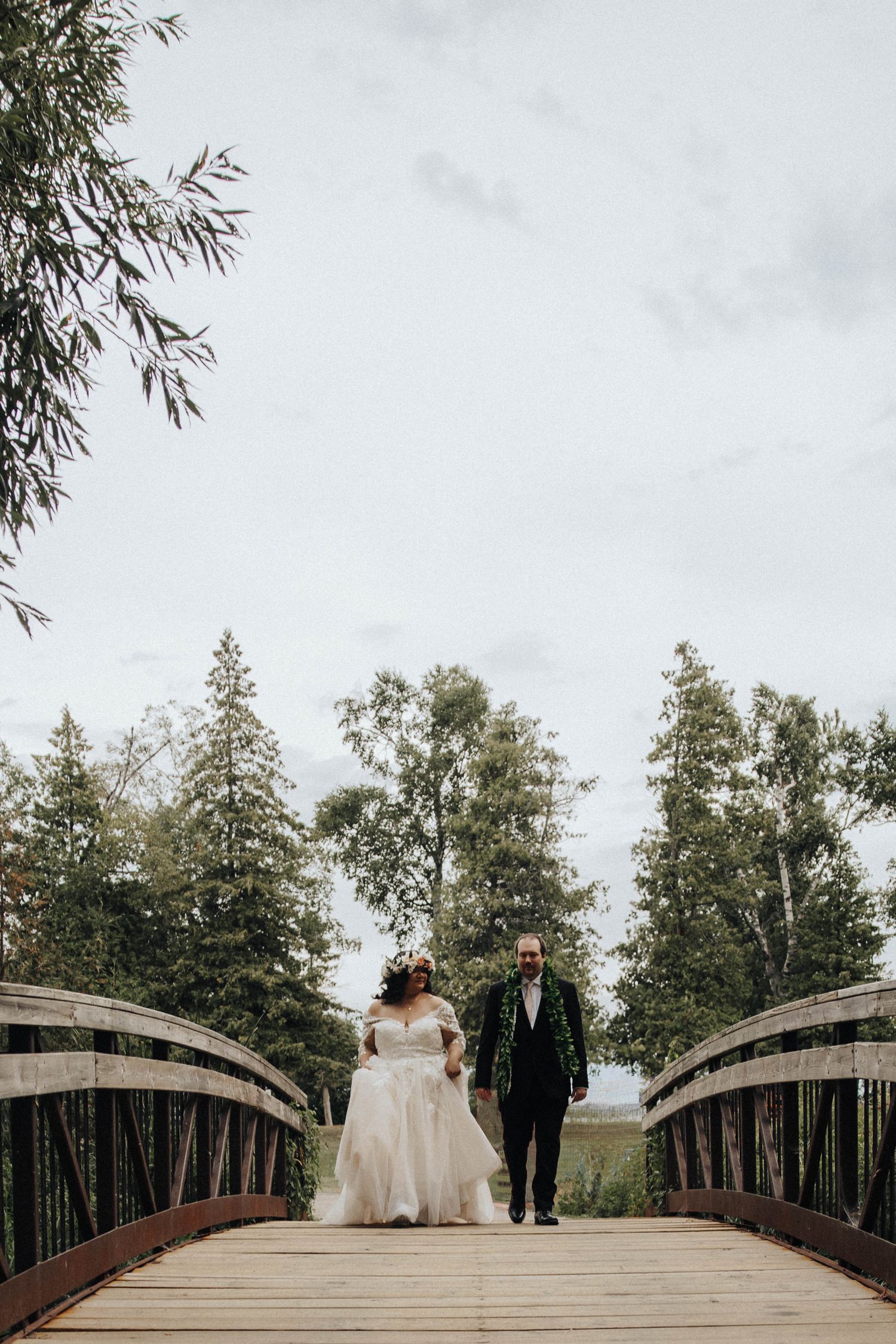Wedding couple walks across rustic wooden bridge surrounded by lush green trees in romantic outdoor setting.