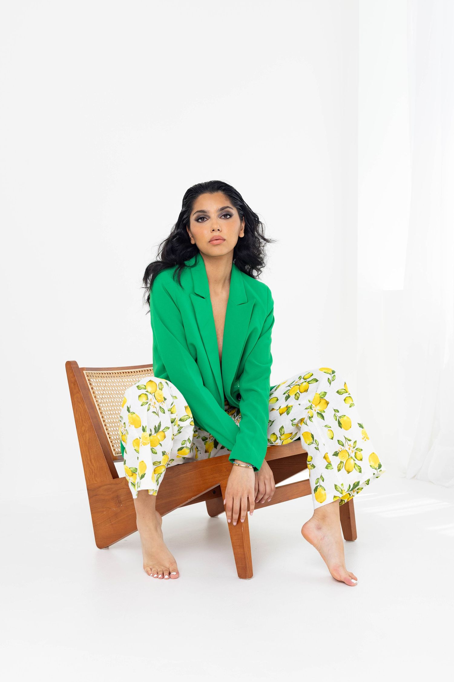 Model in vibrant green blazer and floral pants poses barefoot on wooden chair against white backdrop.