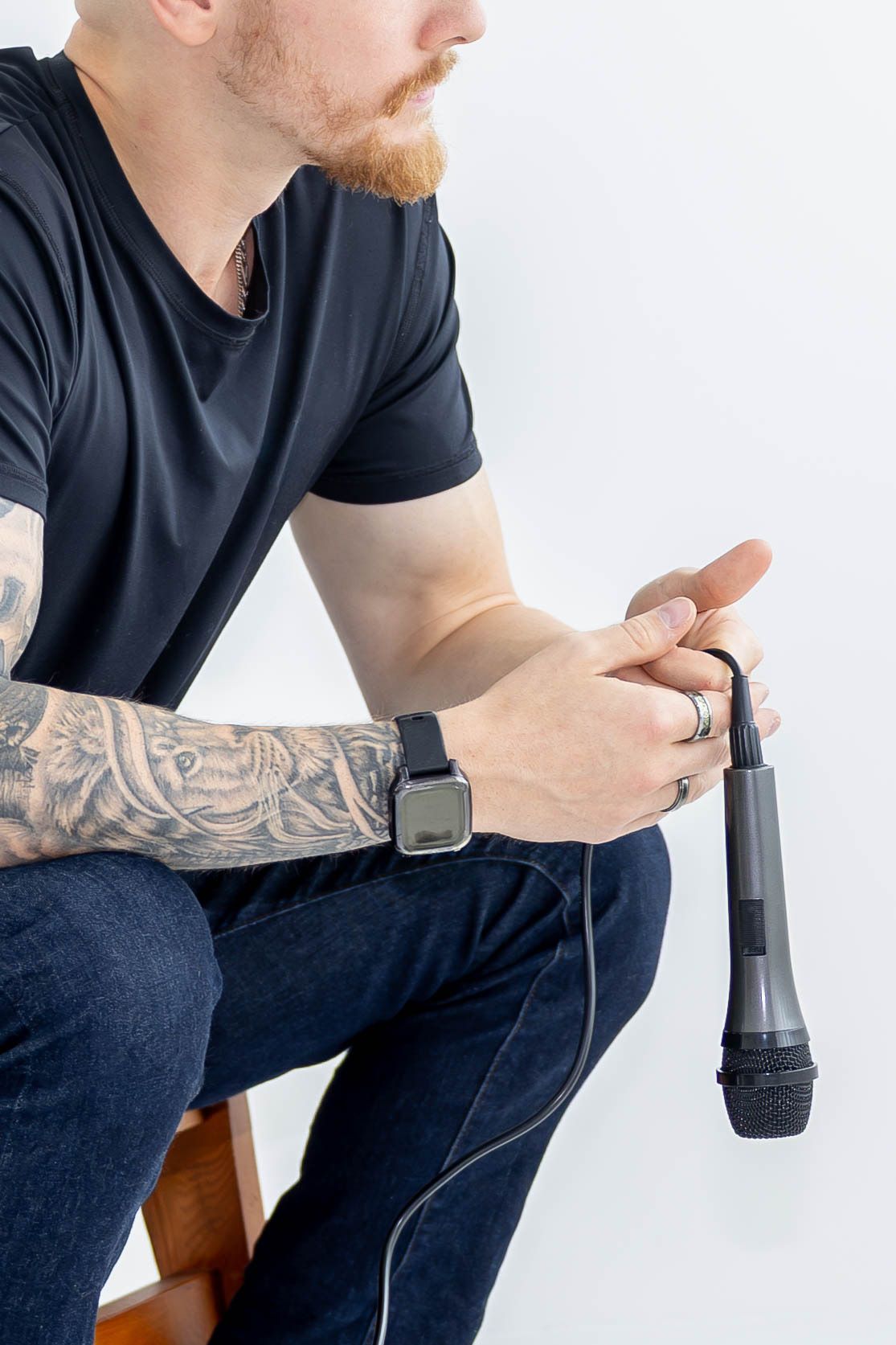Person in black t-shirt and jeans holding a dark device while sitting on wooden furniture against white background.