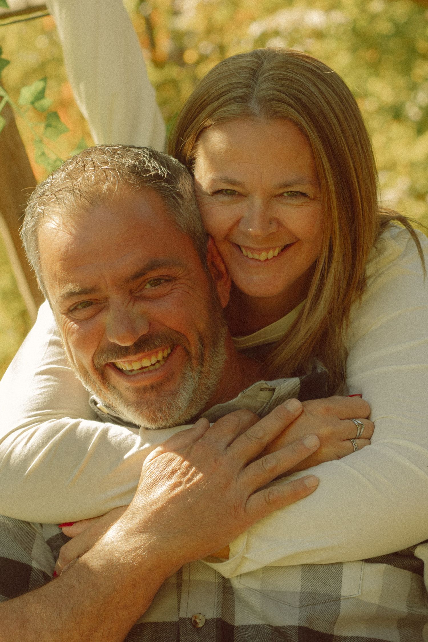 A smiling middle-aged couple embracing outdoors, woman hugging man from behind, both wearing casual cream tops.