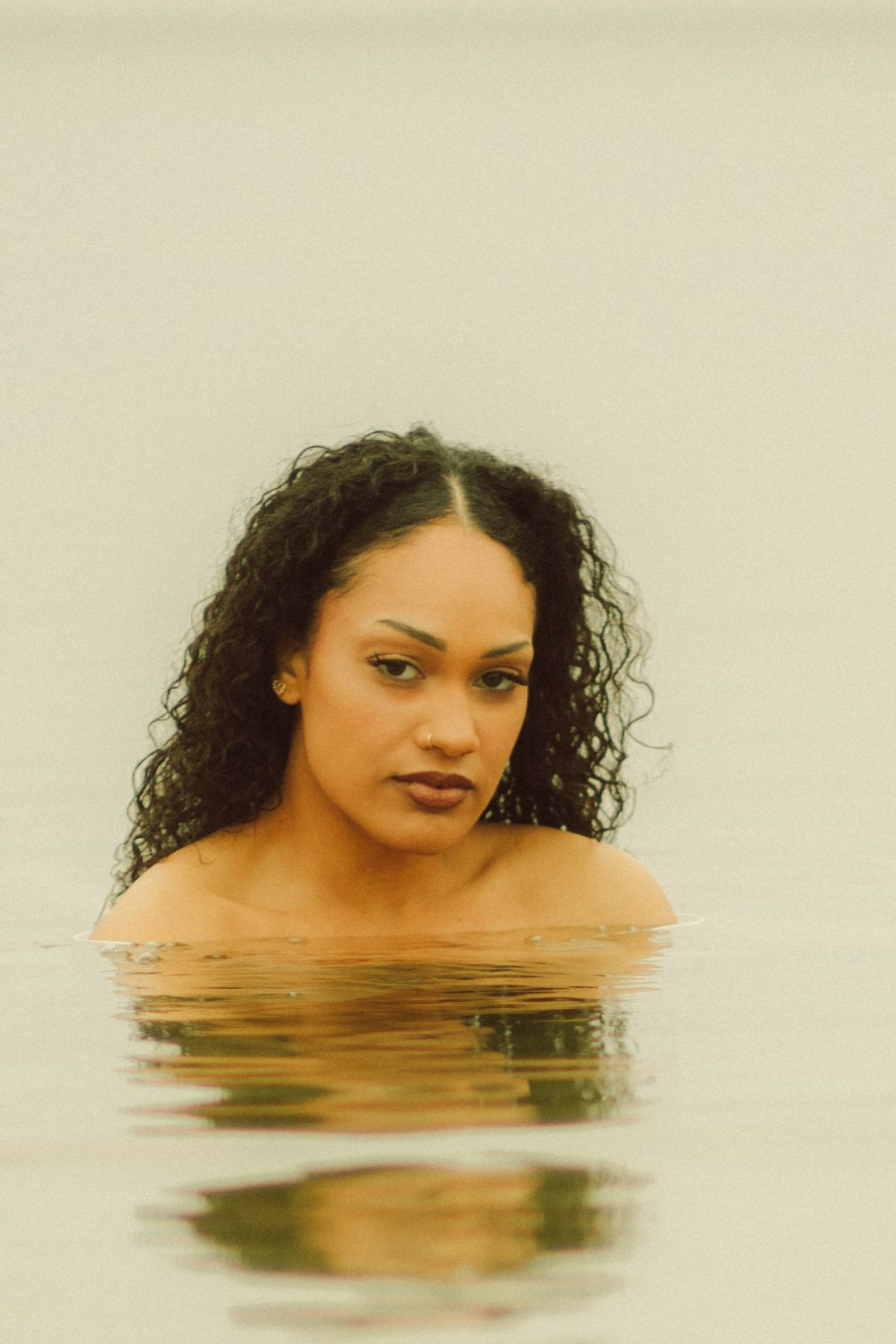A young woman with curly black hair emerges from calm water, her bare shoulders visible above the reflective surface.