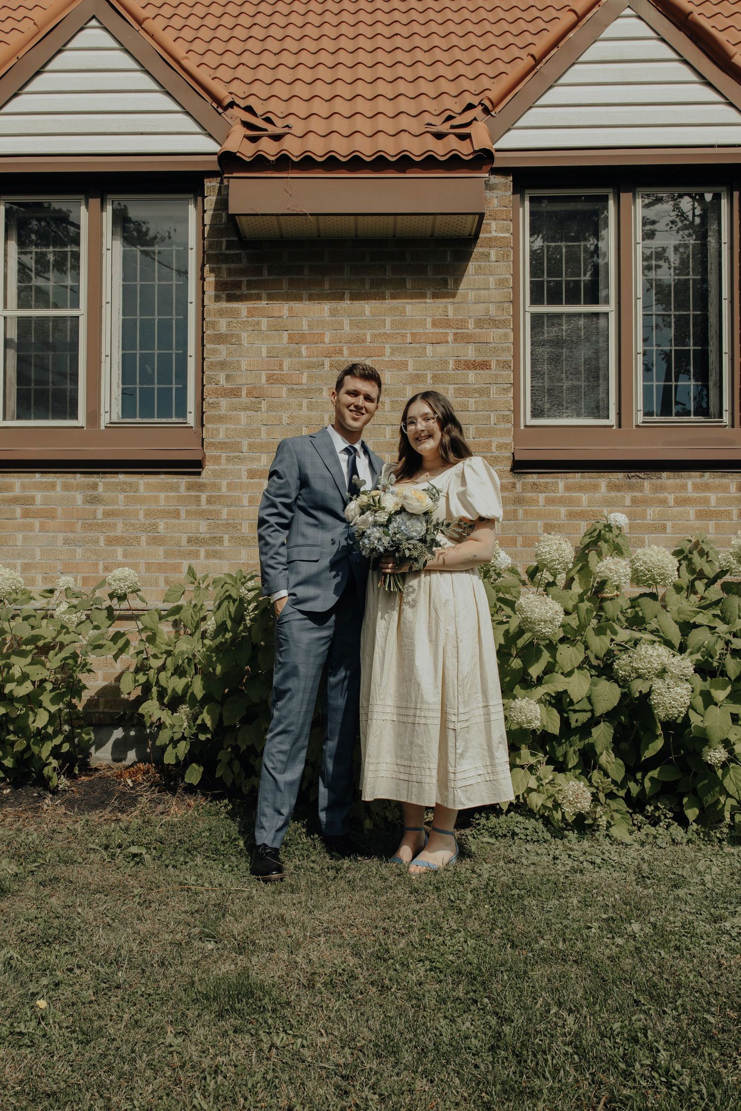 A couple poses by a brick house with terra cotta roof tiles during their wedding day.