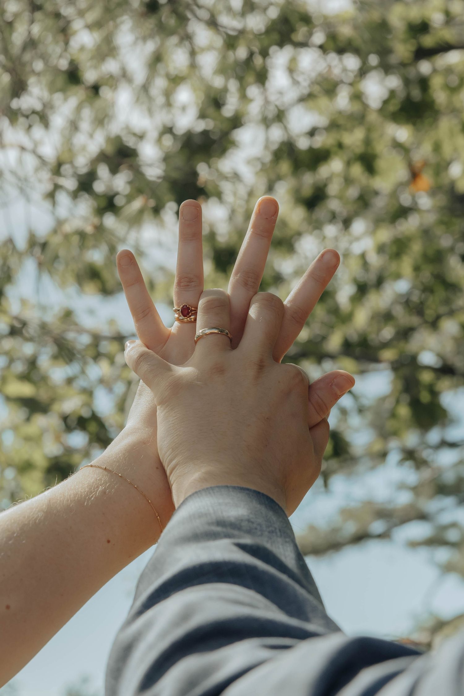 Hands raised against a blue sky showing wedding rings with sunlight filtering through green leaves in the background.