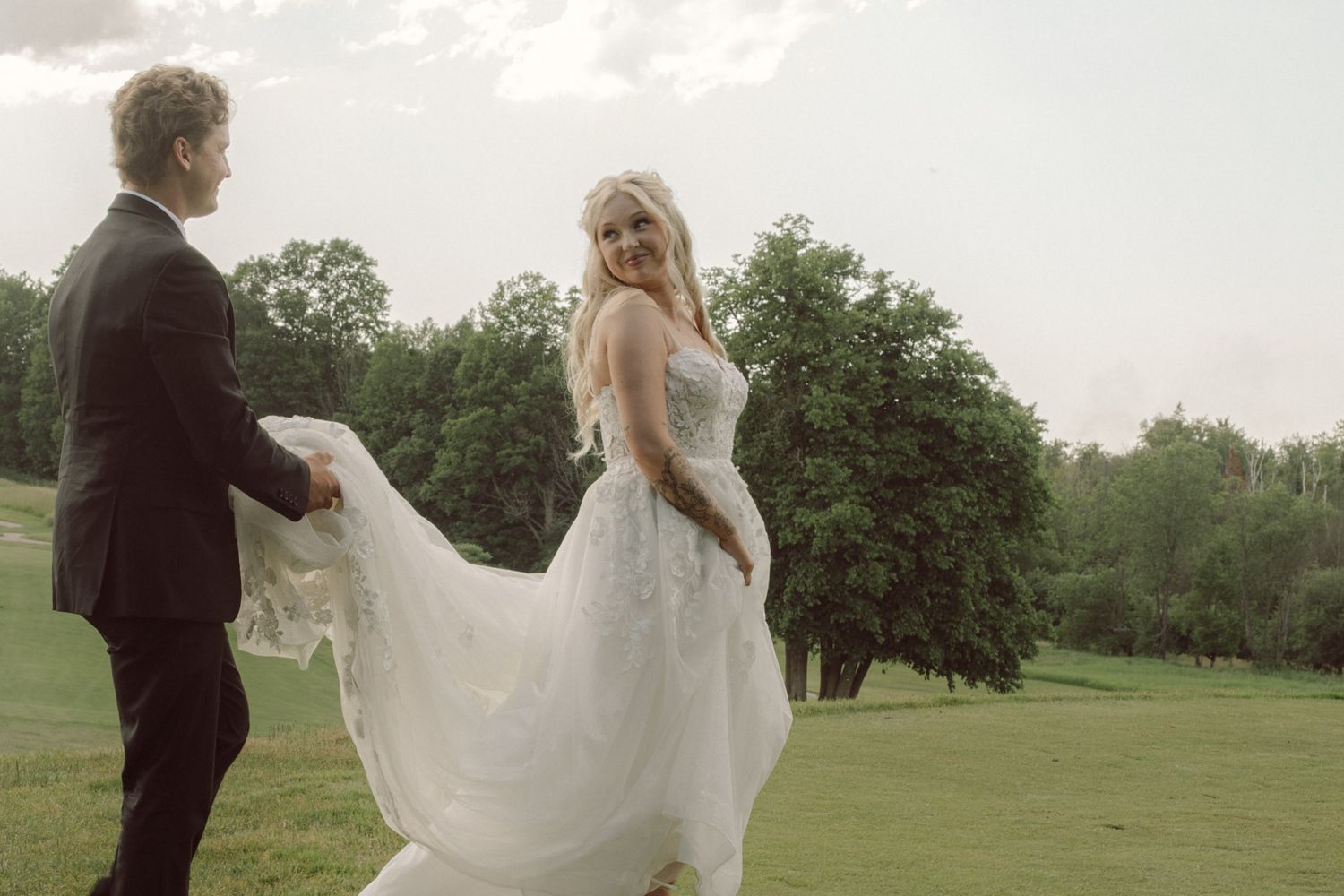 Bride in flowing white gown twirls on green lawn while groom in black suit holds her hand at outdoor wedding.
