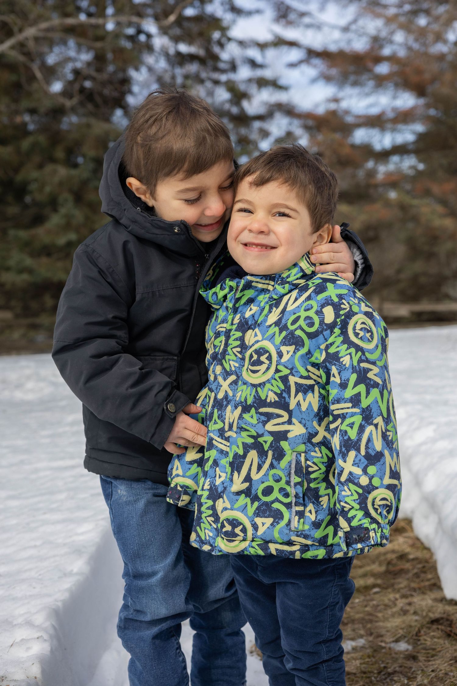 Two children in winter clothing share a playful moment in the snow with one wearing a colorful patterned jacket.