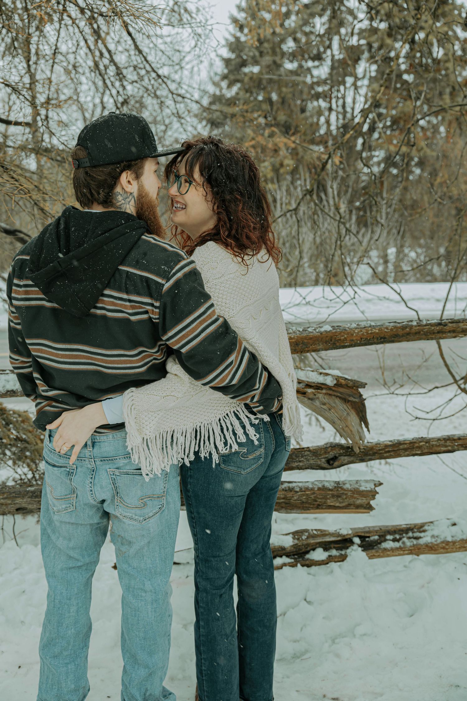 Two people wearing jeans and sweaters embrace while walking in a snowy winter landscape.