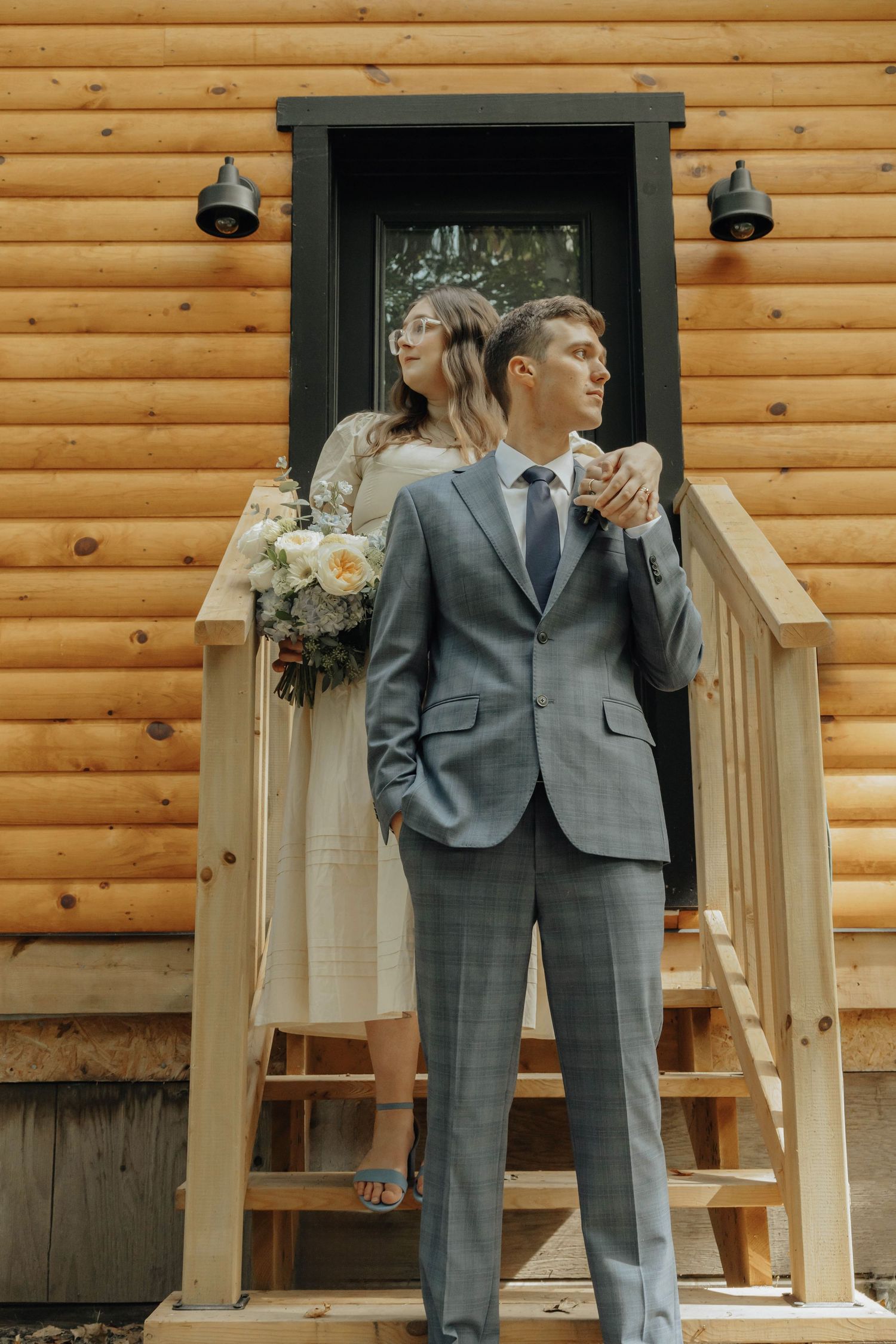 A couple poses in front of a rustic wooden cabin wearing formal wedding attire.
