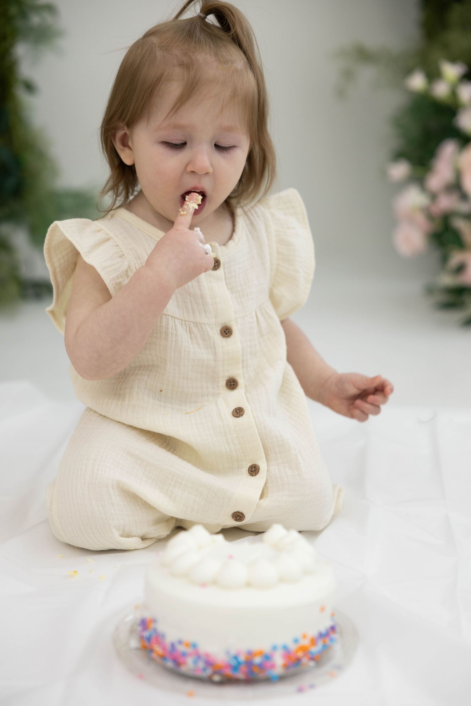 Small toddler in cream button-down romper tasting frosting from a white cake decorated with colorful sprinkles.