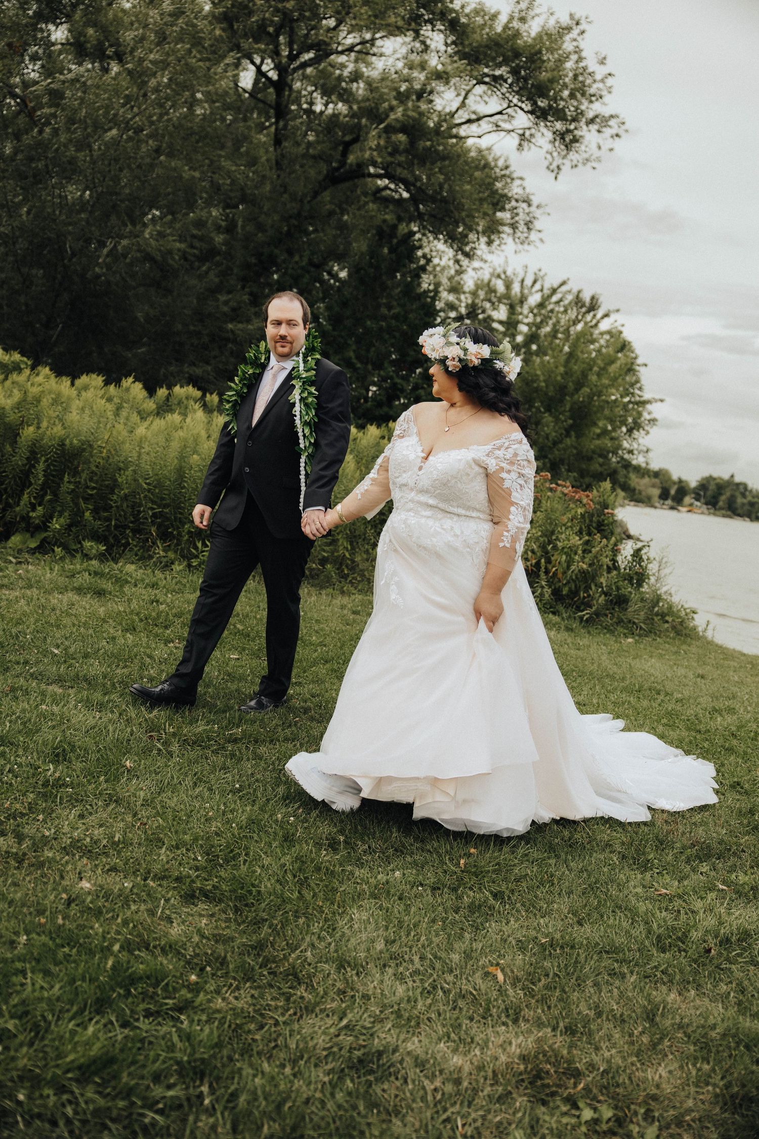 A wedding couple walks together outdoors on a grassy hillside overlooking trees and sky.