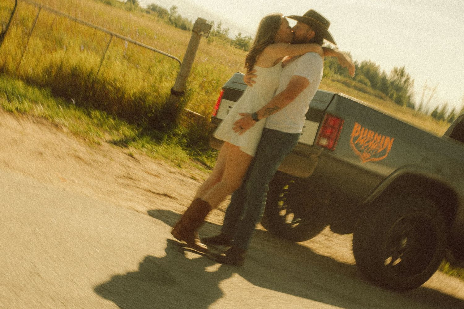 Couple kissing beside a vintage Jeep on a rural dirt road surrounded by open fields.