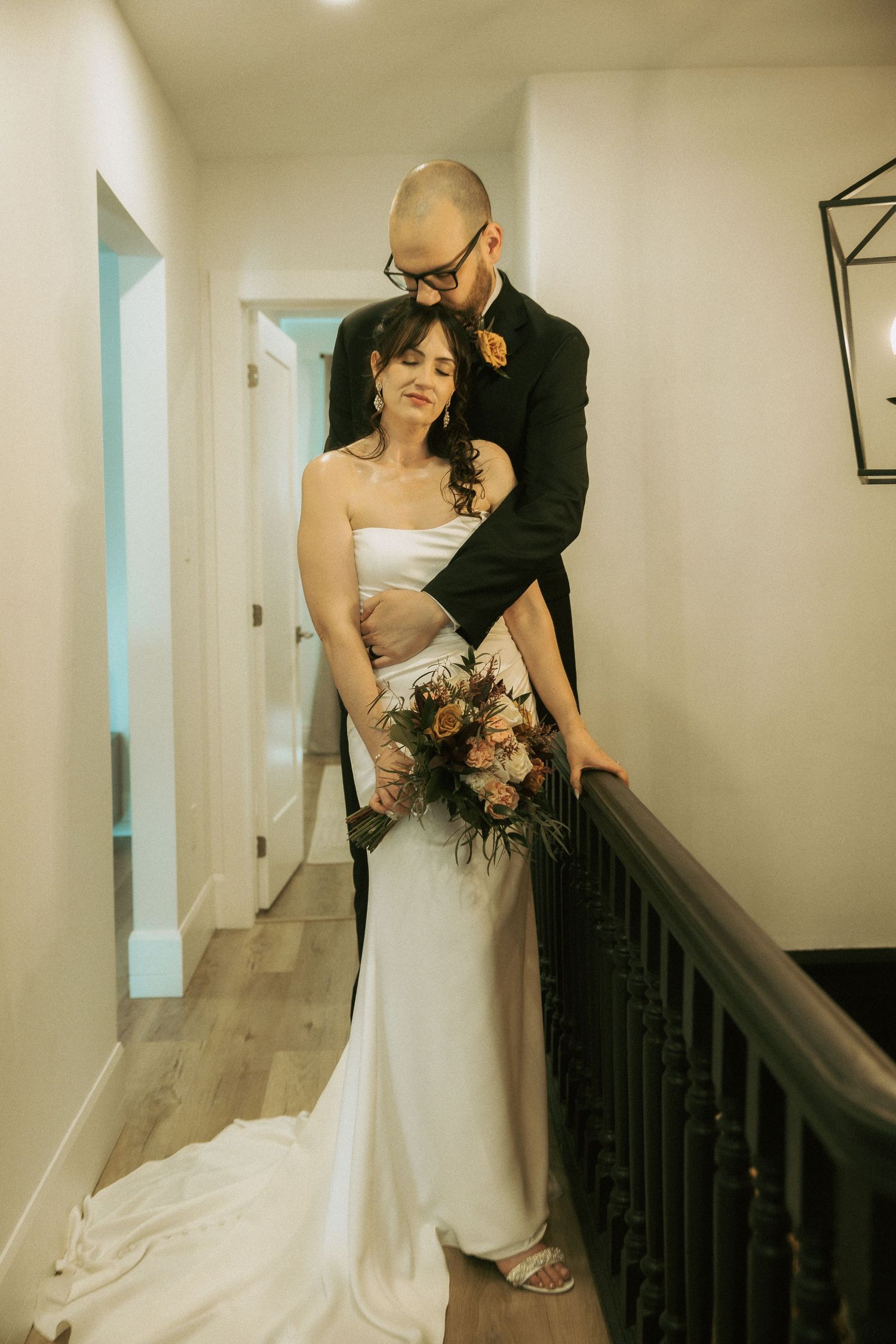 Groom embraces bride in white gown on hallway balcony, both looking down tenderly.