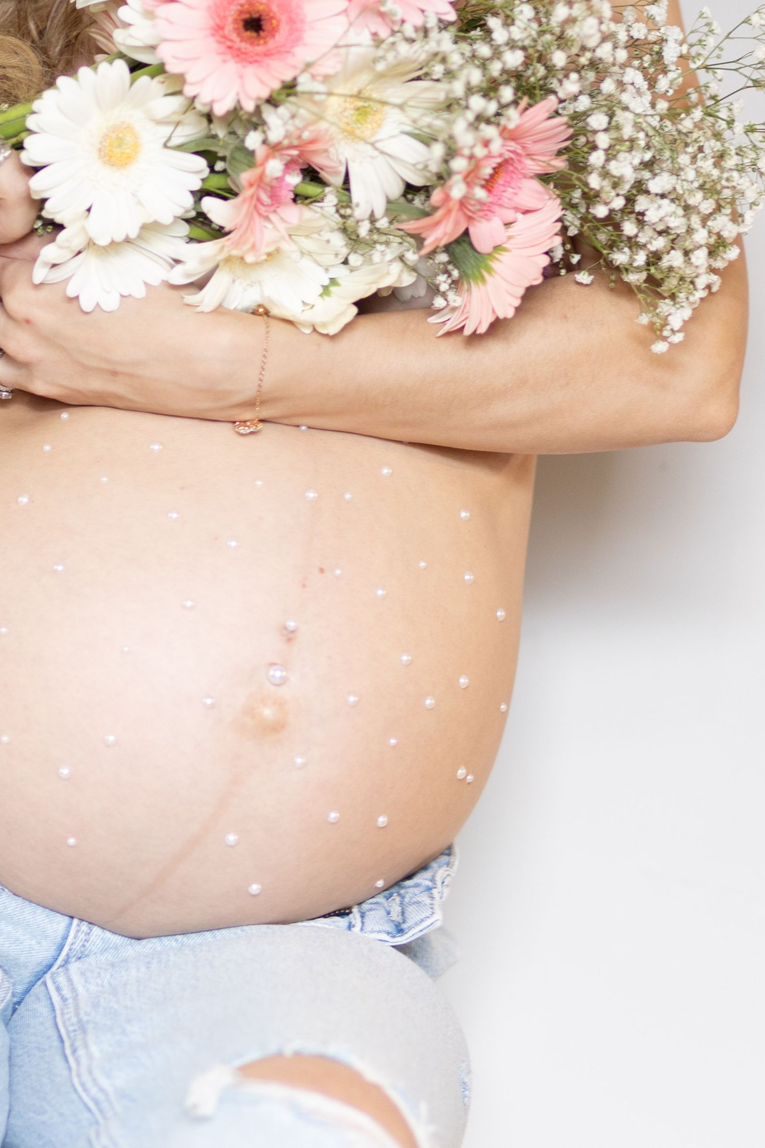 Close-up of pregnant belly in ripped jeans adorned with pearl dots, holding a bouquet of pink flowers.