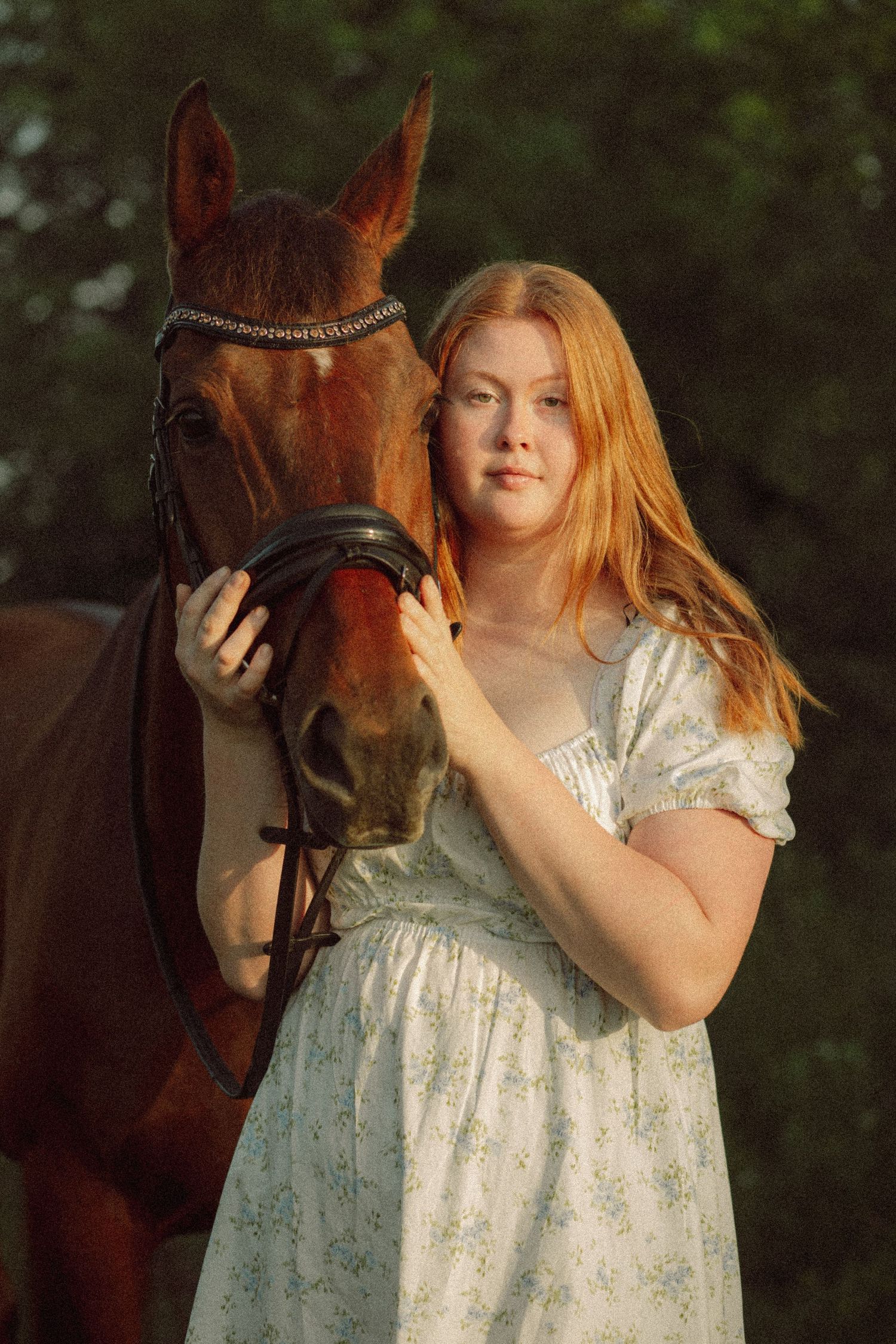Young redhead woman in floral dress posing with chestnut horse outdoors in golden hour light.