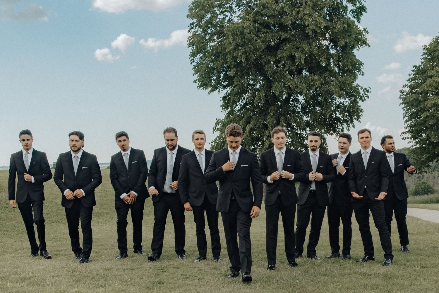 Groom and groomsmen in black suits posing outdoors on green lawn with large tree backdrop at wedding.