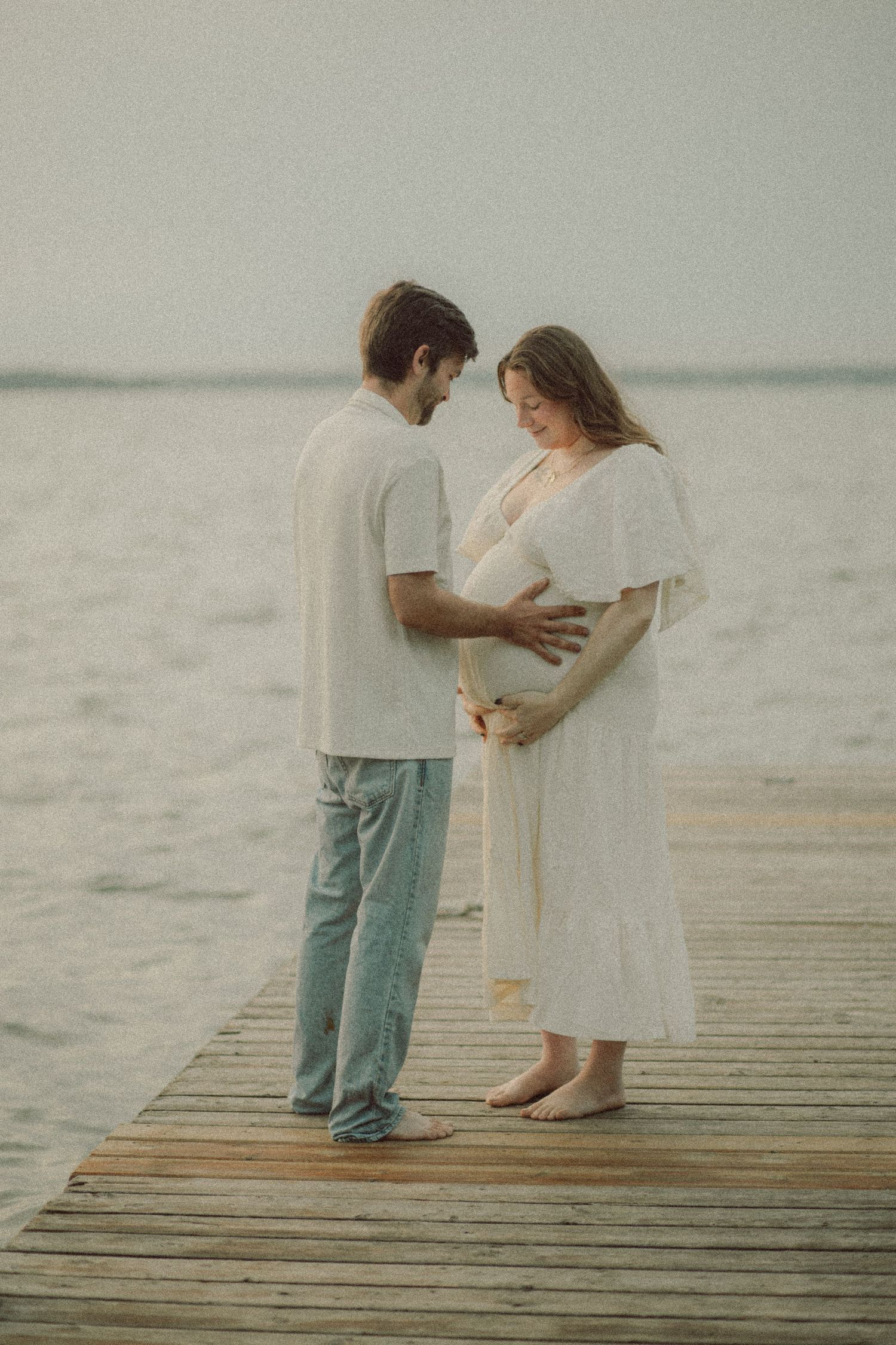 Couple sharing a tender moment on a wooden dock by the water, woman in white dress cradling her pregnant belly.