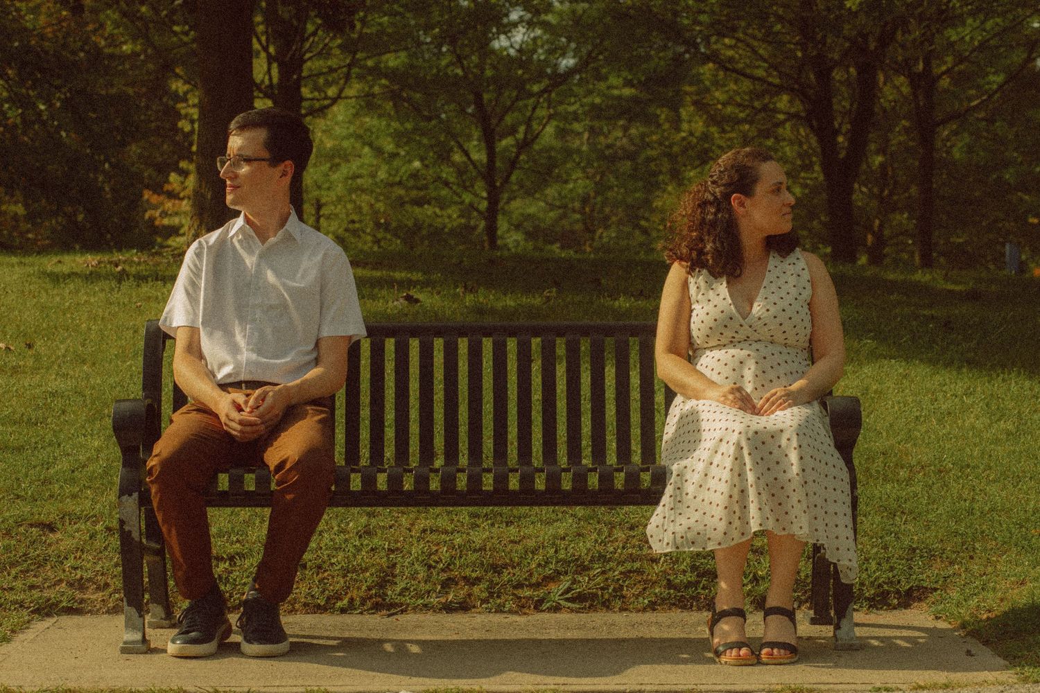Two people sitting apart on a park bench outdoors, surrounded by green trees on a sunny day.