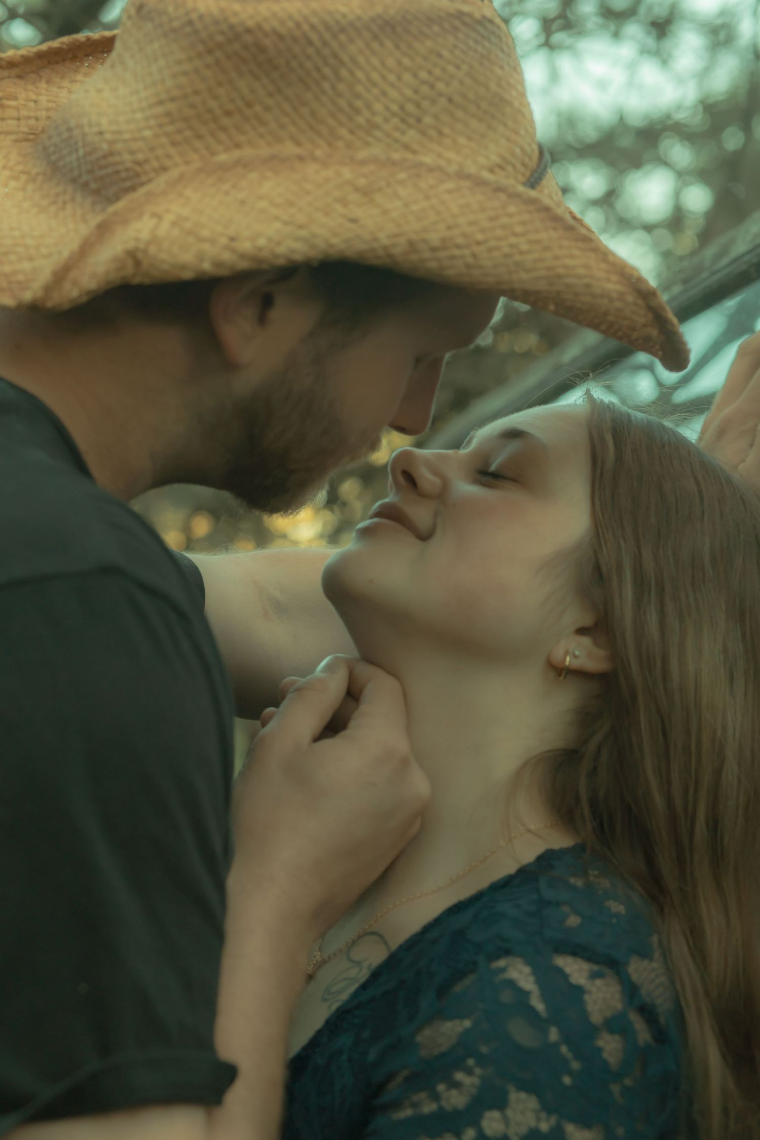 Bearded man in straw cowboy hat leaning in to kiss smiling woman in blue lace top outdoors.