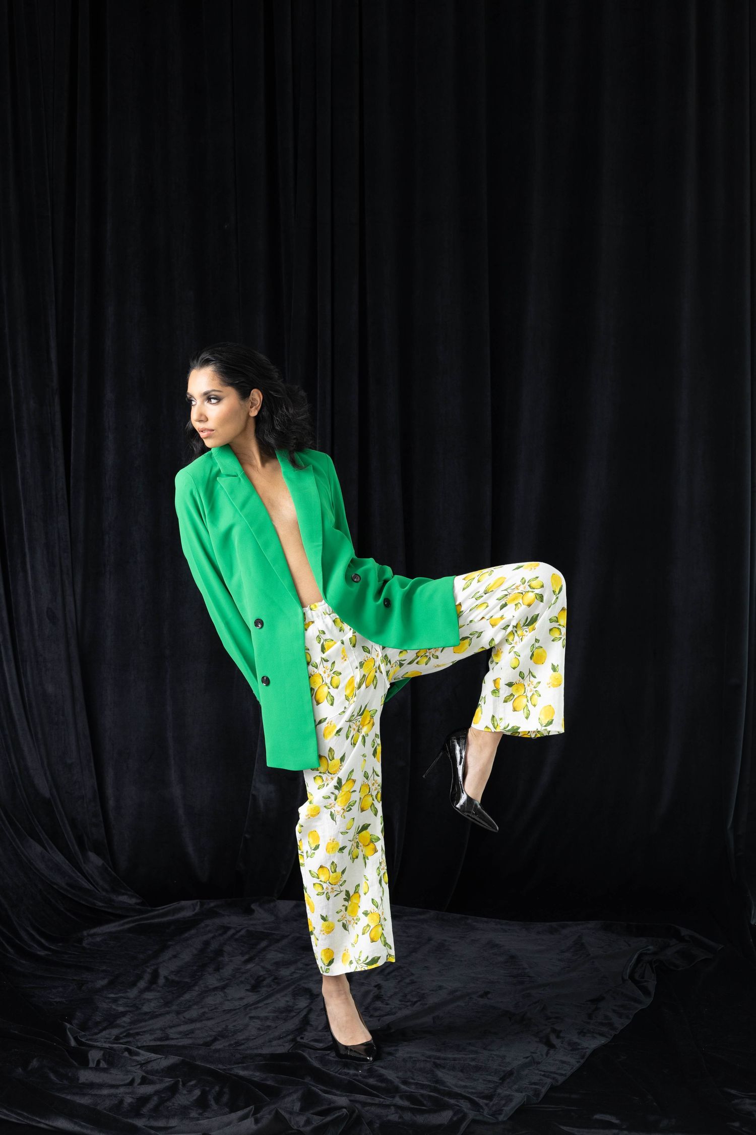 Model in bright green blazer and floral dress poses dramatically against black backdrop in fashion photo series.