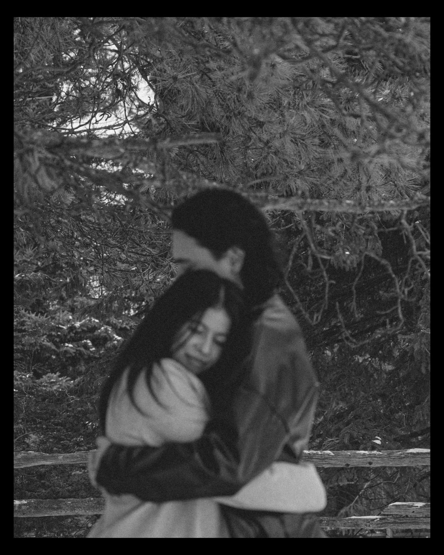 A black and white photograph shows two people embracing outdoors with tree branches visible in the background.