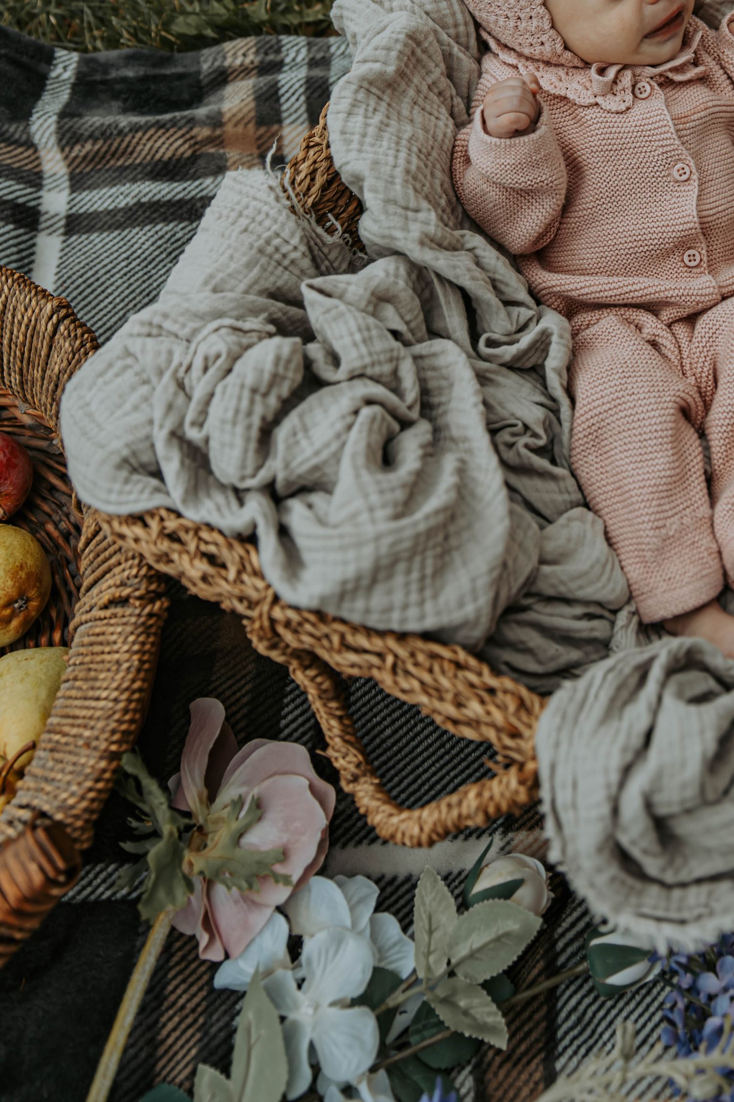 Cozy pink knit sweater and gray blankets arranged in wicker basket with delicate flowers on plaid background.