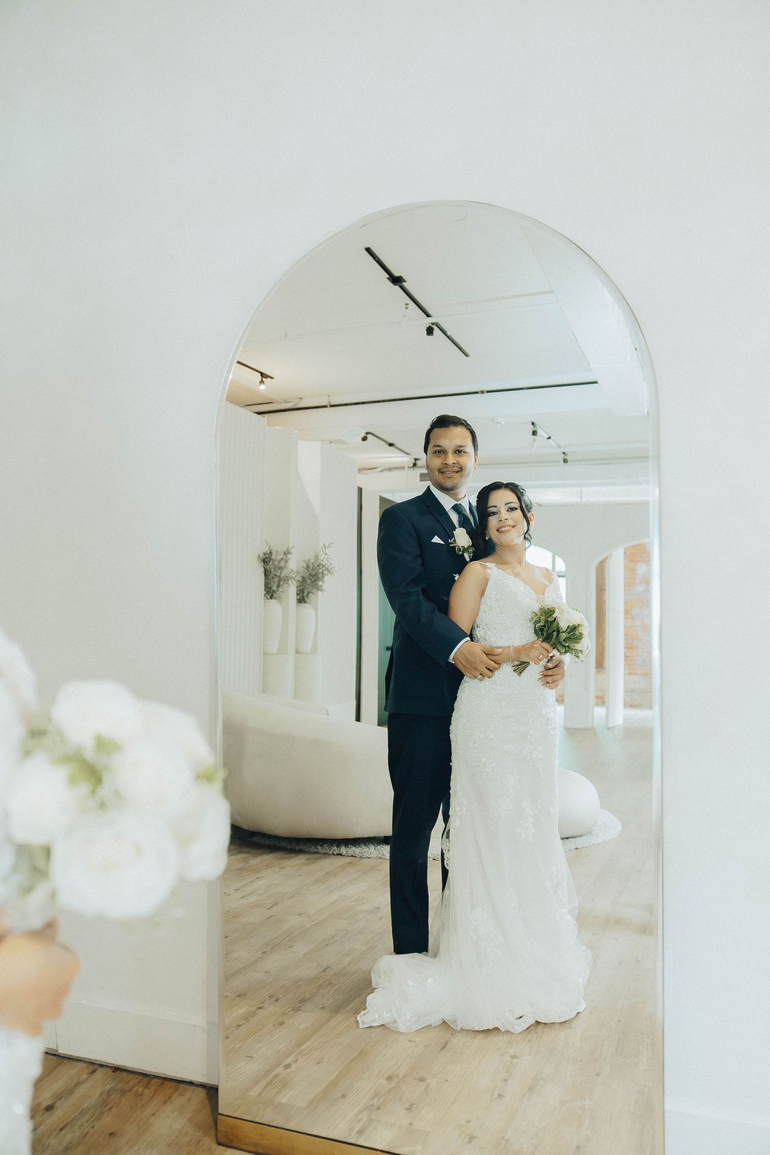A couple in wedding attire stands in an elegant white room with arched doorways and light wood flooring.
