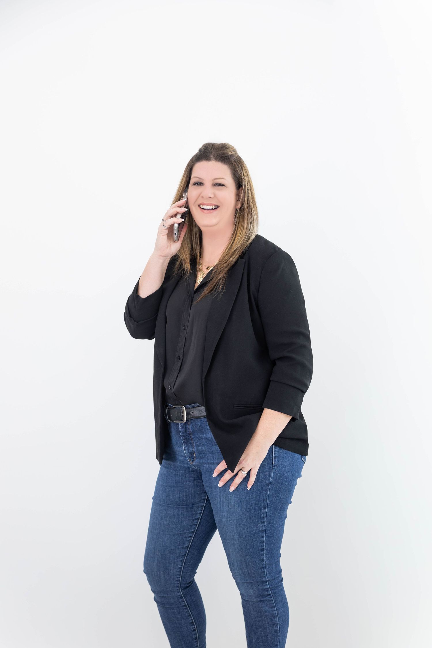 A person wearing a black blazer and jeans laughs while talking on a phone against a white background.