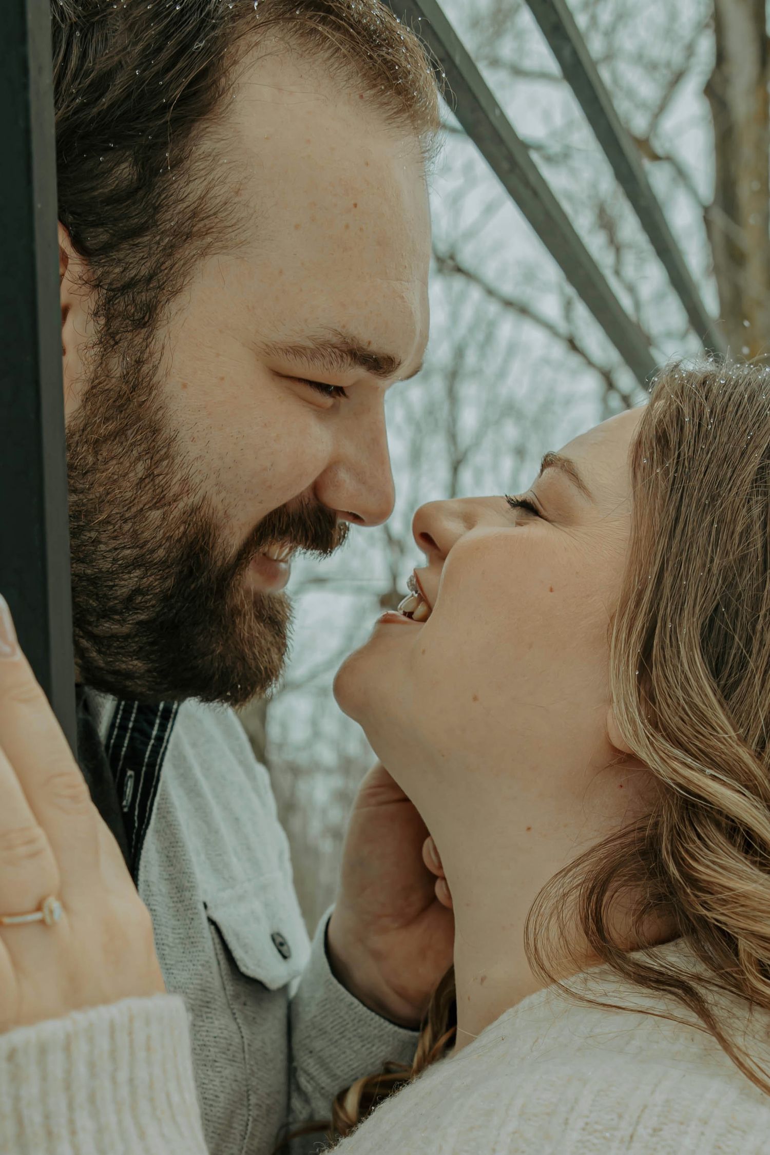 A romantic close-up moment between a couple in winter, with him sporting a beard and her with wavy blonde hair.