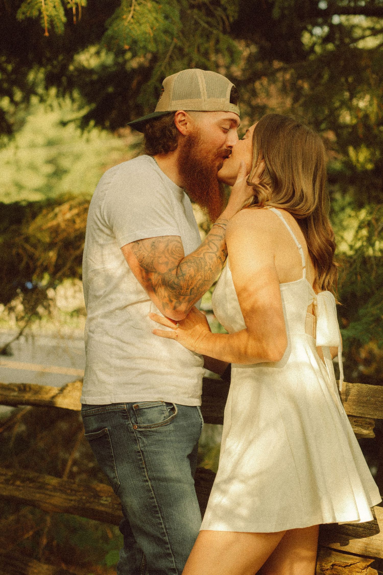 Bearded tattooed man in cap kissing woman in white dress outdoors near wooden fence with green trees.