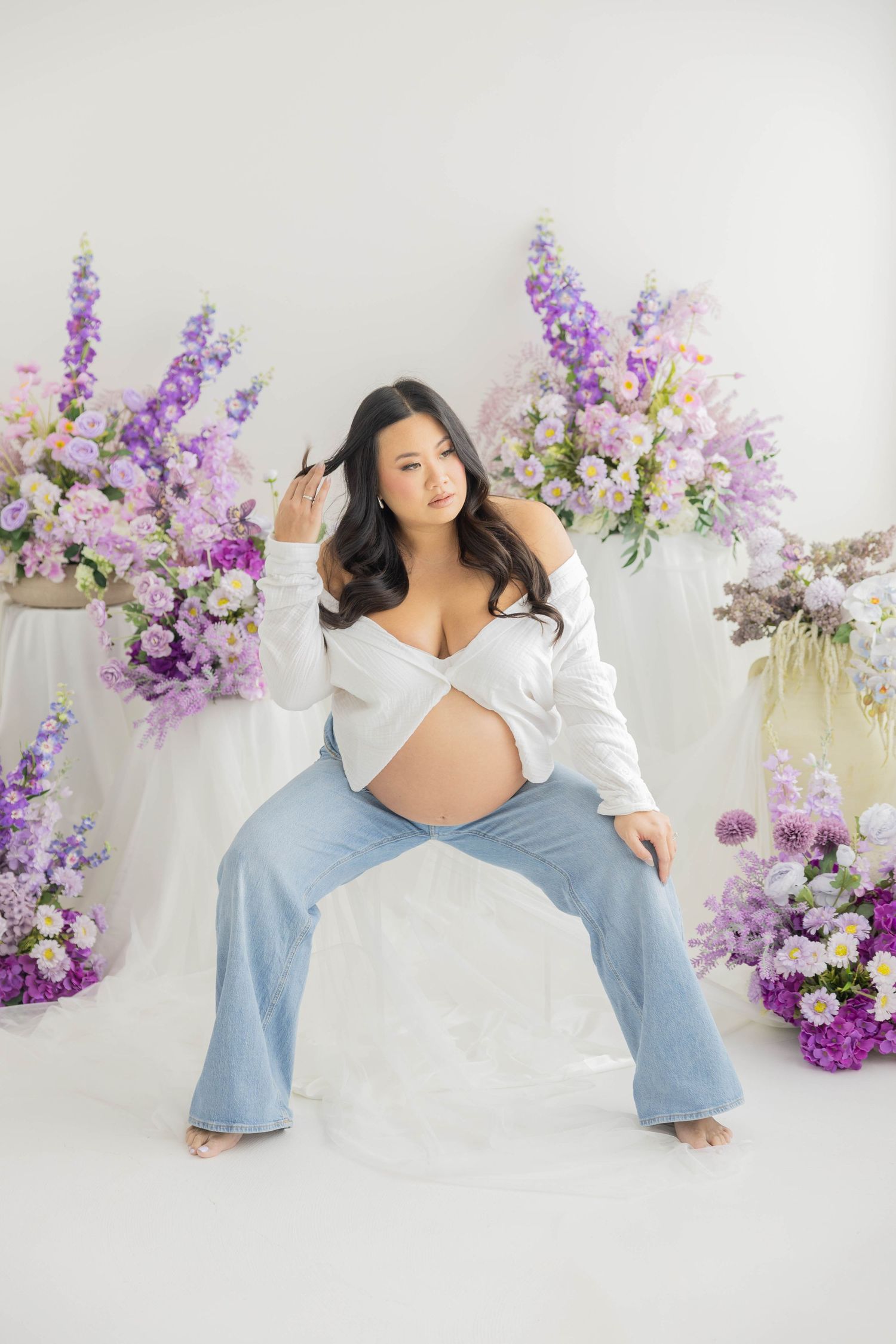 A woman poses gracefully in white and denim against a backdrop of purple delphinium flowers in a bright studio setting.