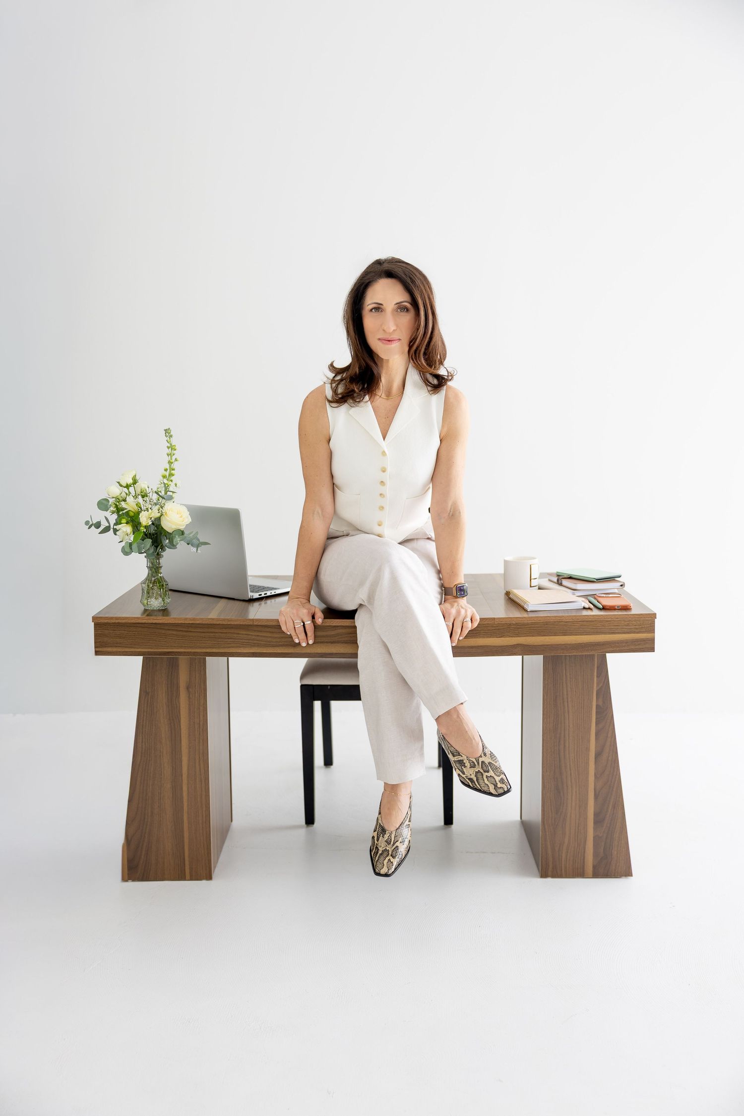 Professional woman in white outfit sitting on wooden desk with laptop and flowers in modern office.