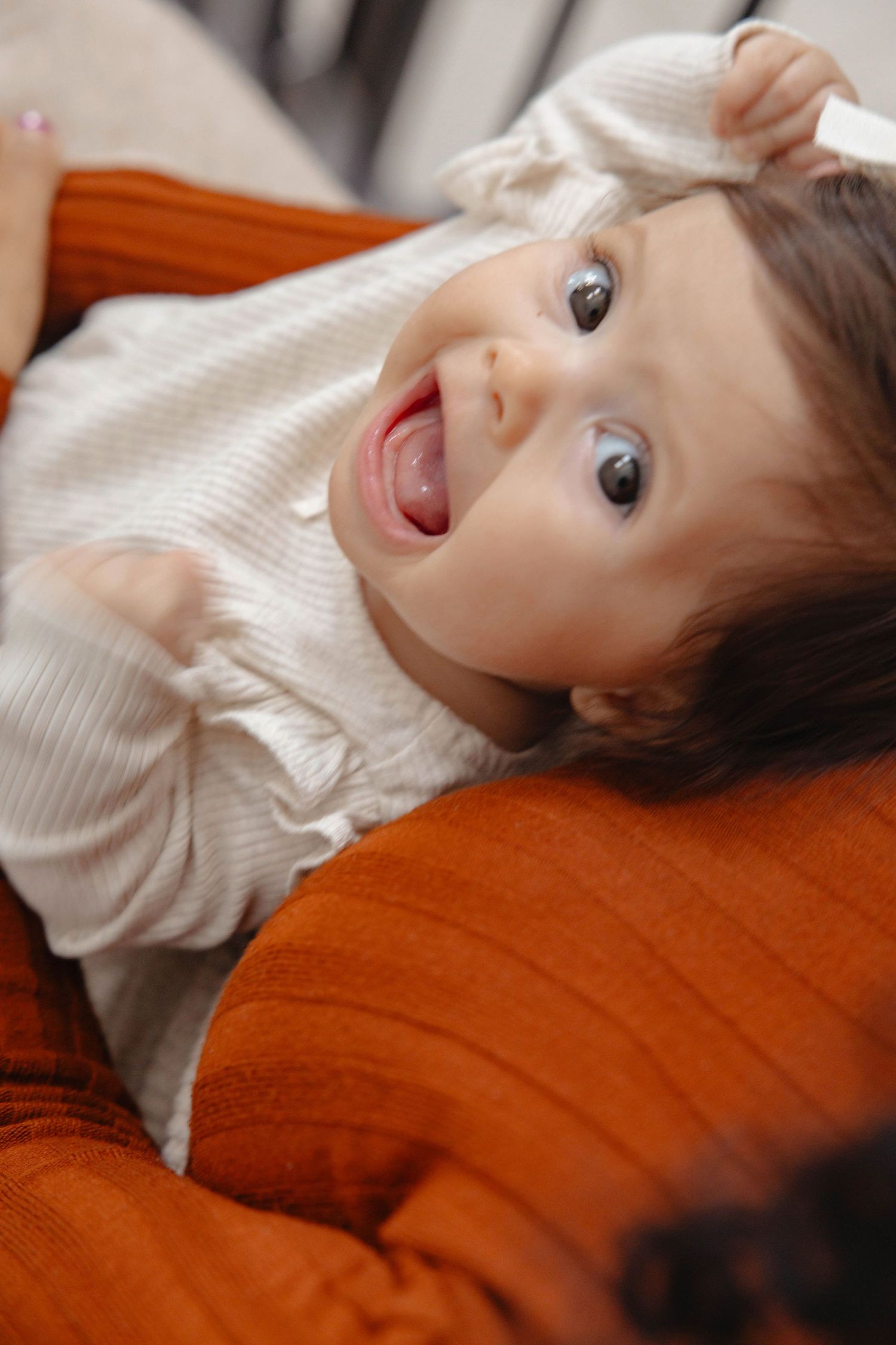 A joyful baby in a white sweater lies on an orange cushion with a bright smile and sparkling blue eyes.